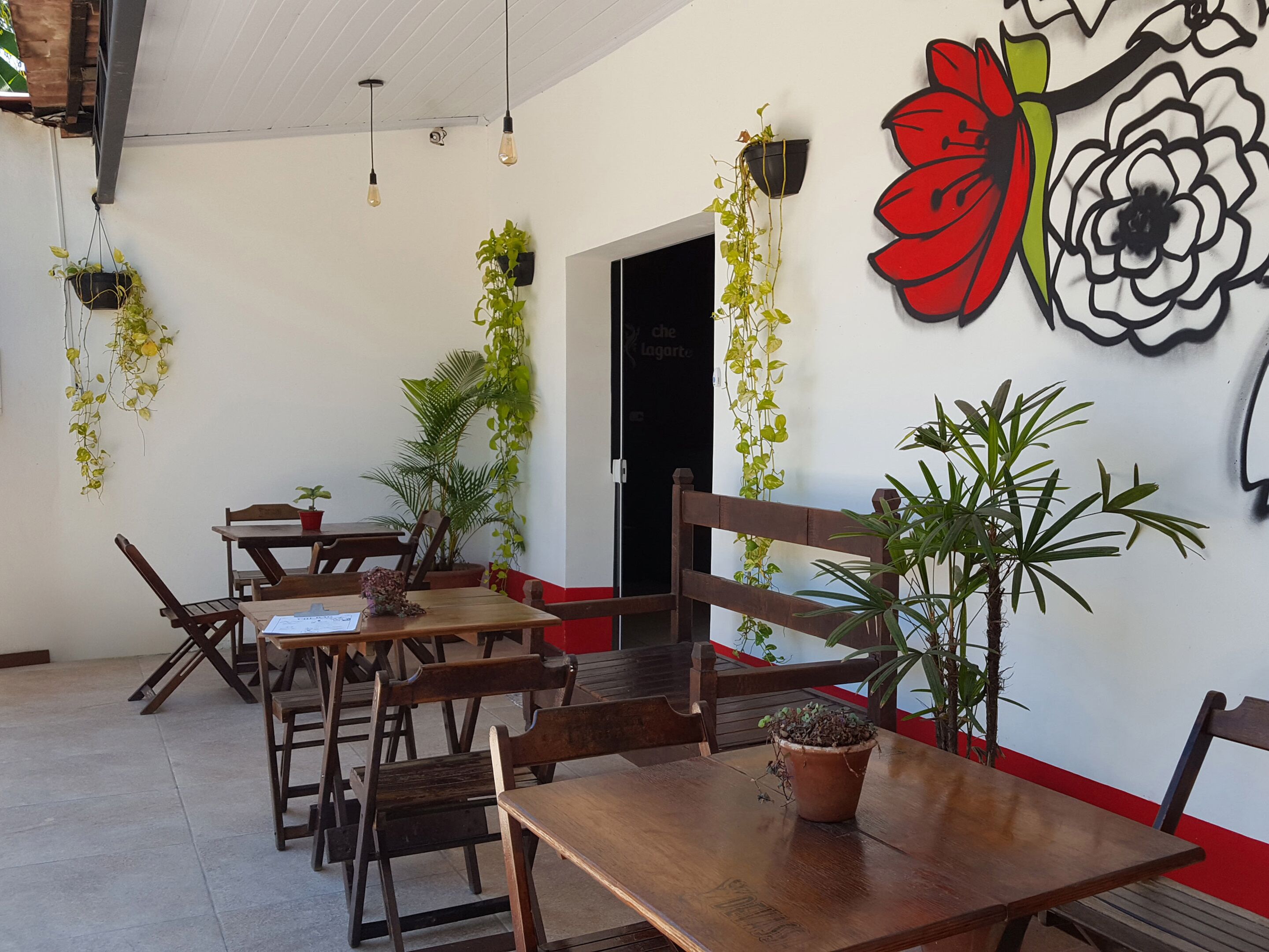 A dining area with wooden tables and chairs, hanging plants, and a wall with a floral mural.