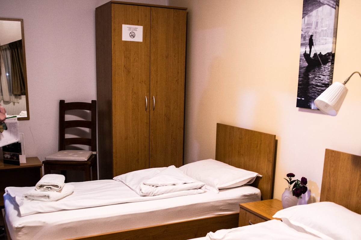 Habitación de hotel con dos camas individuales, un armario de madera, un escritorio con silla, un espejo y un cartel en la pared.