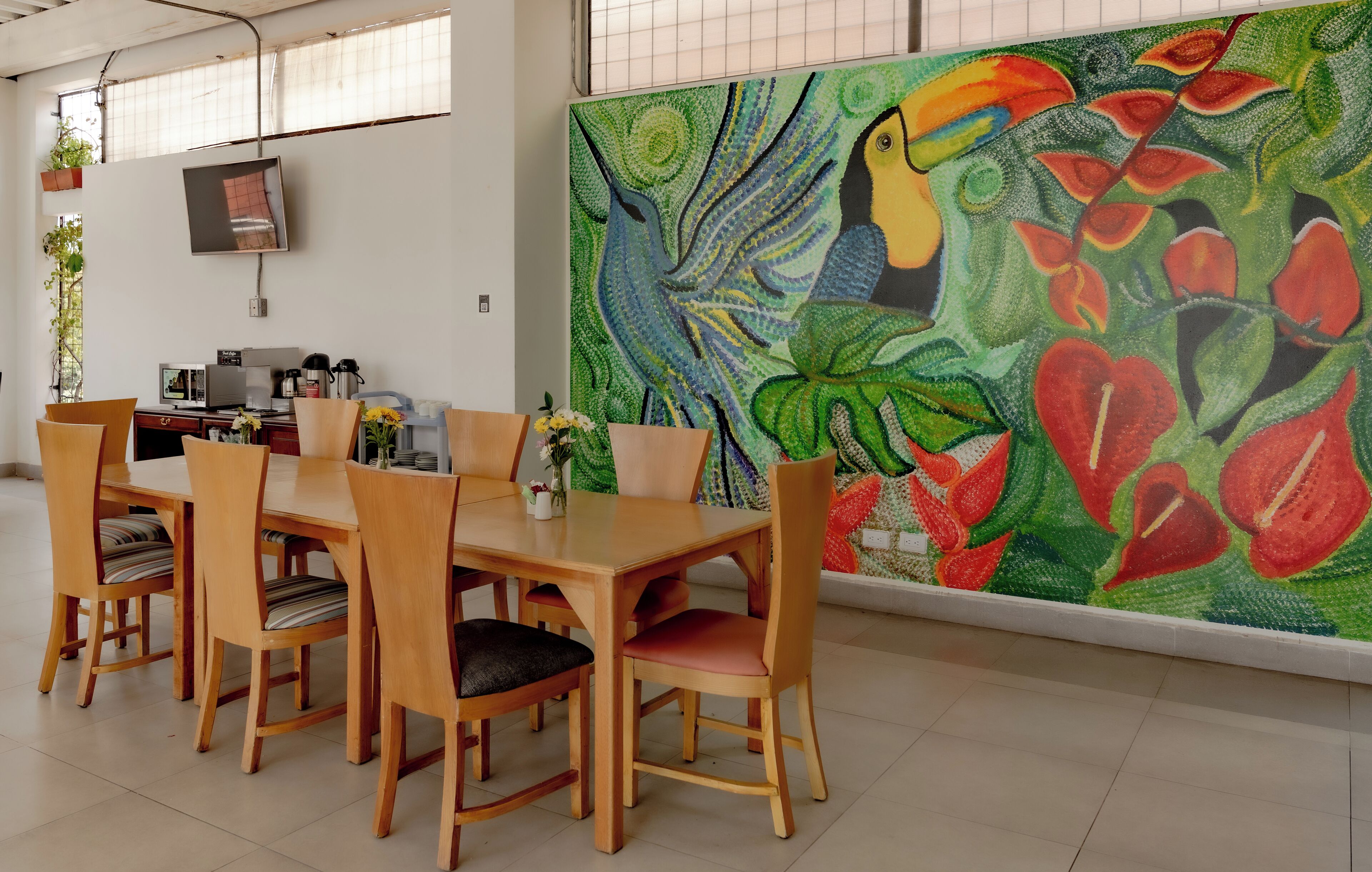 A dining area with a wooden table and chairs, a TV mounted on the wall, and a colorful abstract painting on the wall.