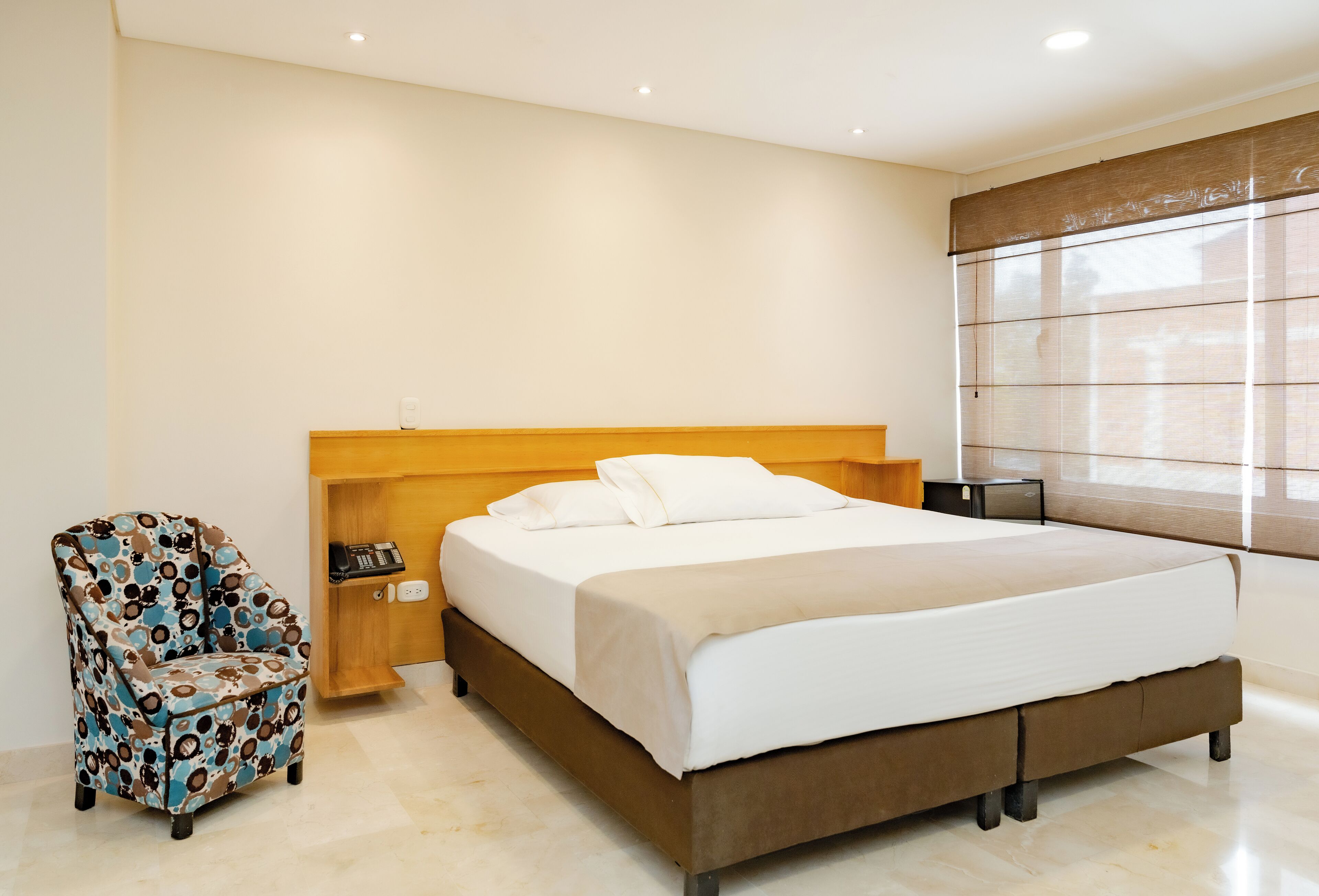 A neatly arranged bedroom with a bed, a patterned armchair, a bedside table with a phone, and a window with blinds.