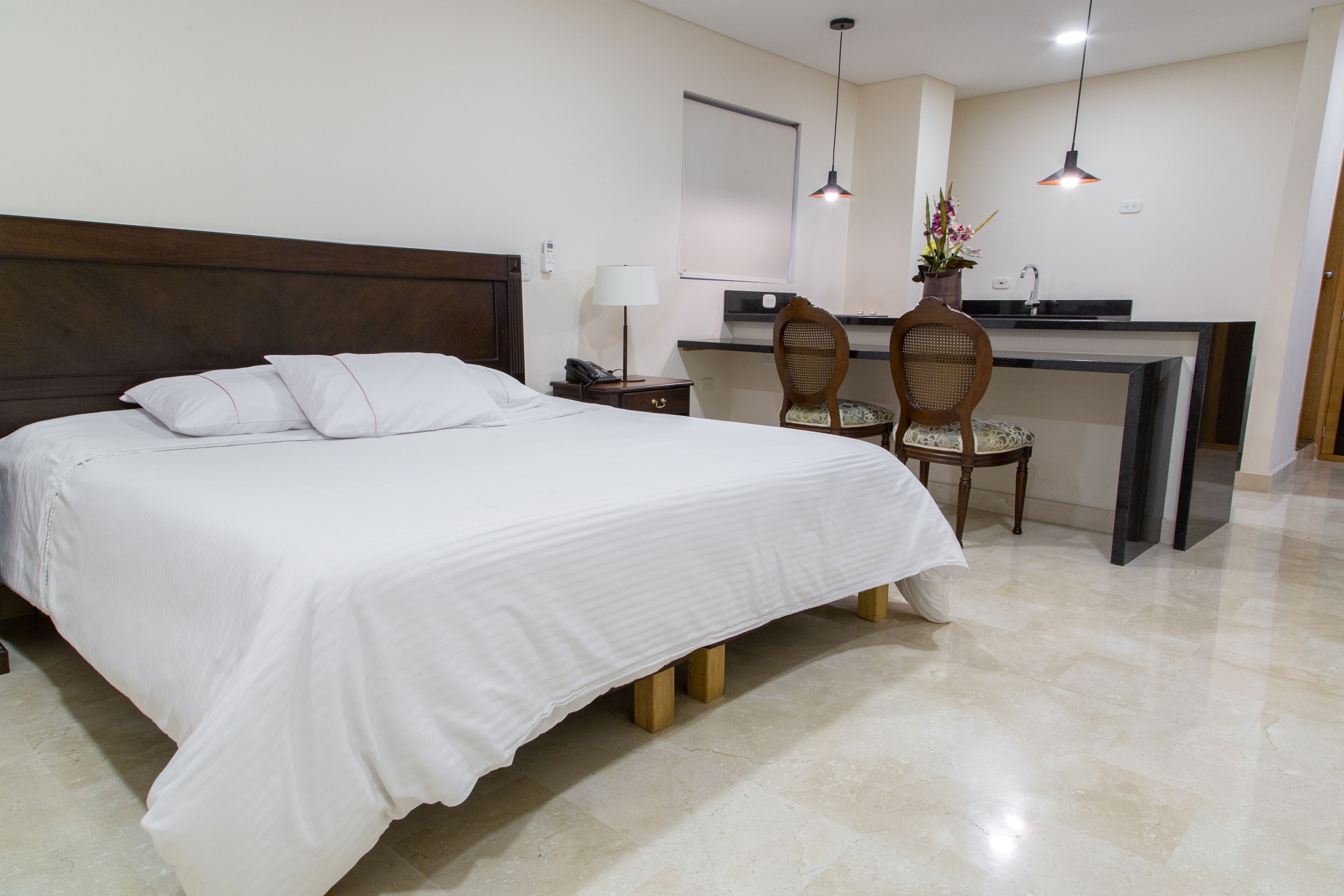 A hotel room with a bed, a desk, two chairs, a lamp, and a potted plant.