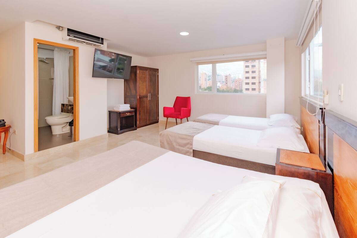 A hotel room with two beds, a TV, a red chair, and a view of the city through the window.