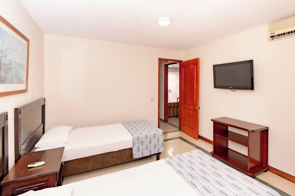 A hotel room with a single bed, a bedside table with a remote control, a TV mounted on the wall, and a wooden side table.