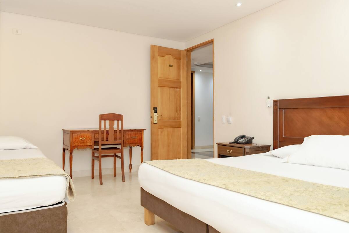 A hotel room with two beds, a desk with a chair, a wooden door, and a bedside table with a phone.