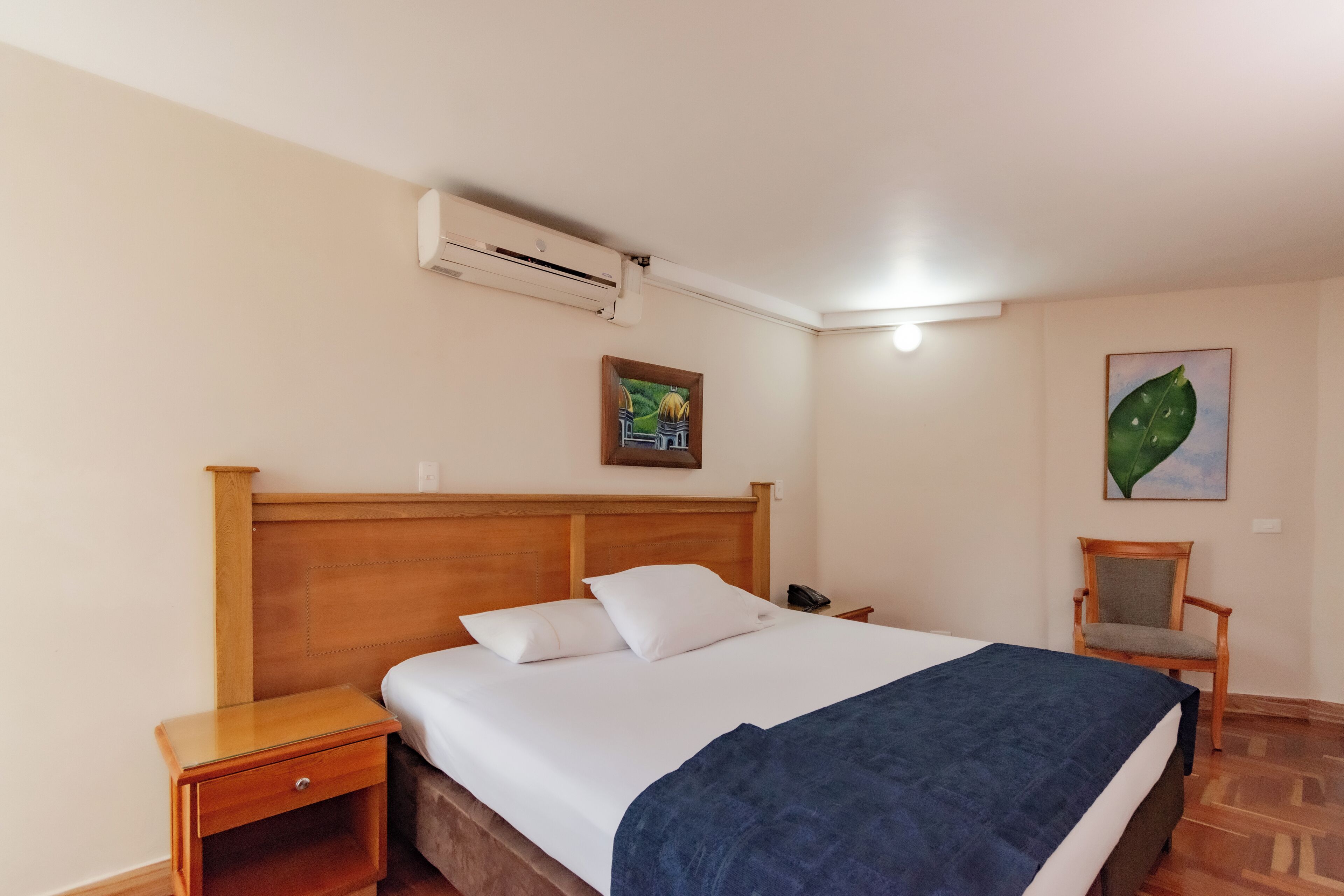 A neatly made bed with a blue blanket, a wooden headboard, a nightstand with a drawer, a chair, and two wall-mounted paintings.