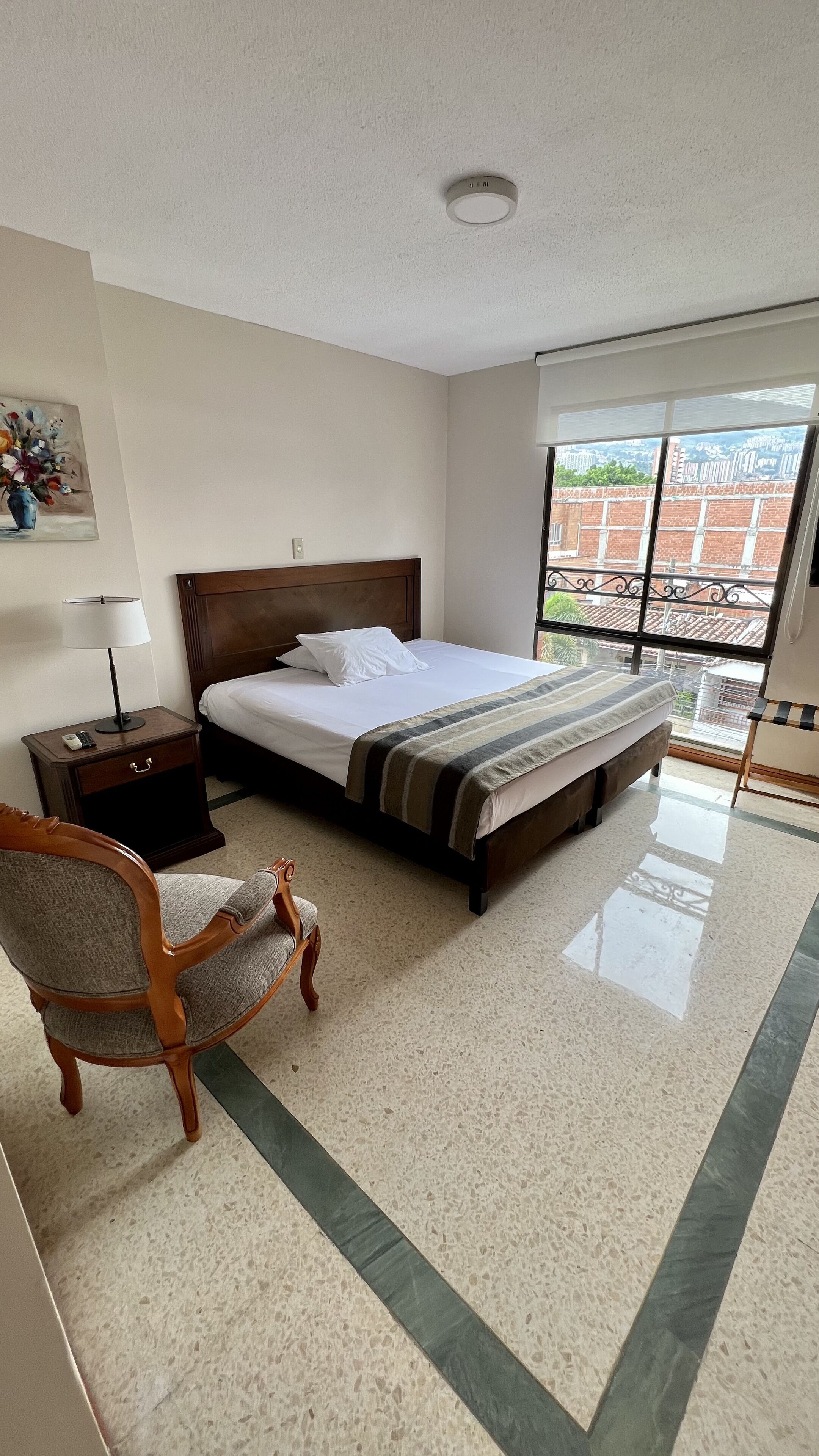 Habitación de hotel con cama, una silla, mesita de noche, lámpara y ventana con vistas a edificios.