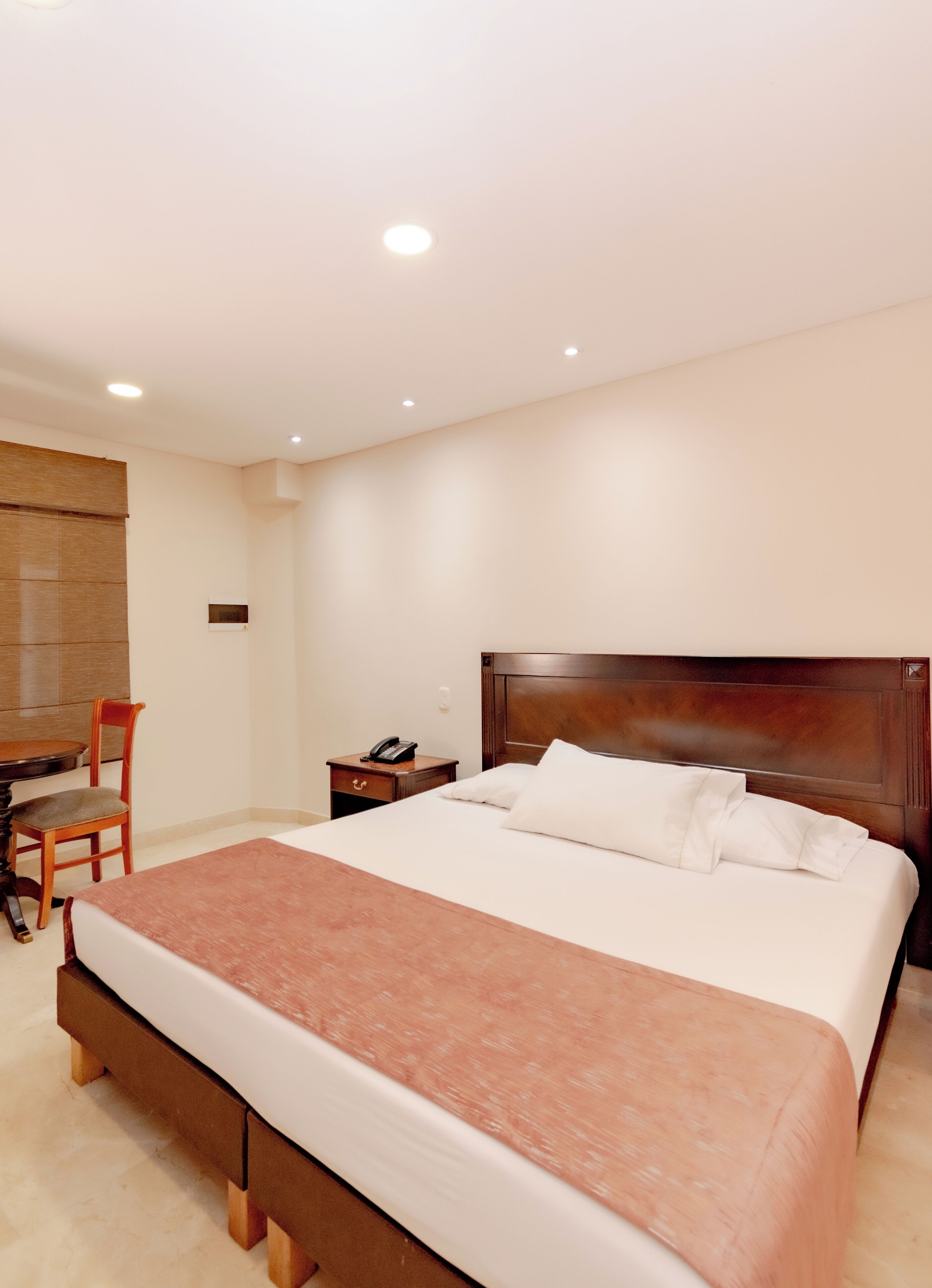 A hotel room with a large bed, a wooden nightstand, a chair, a desk, and a telephone.