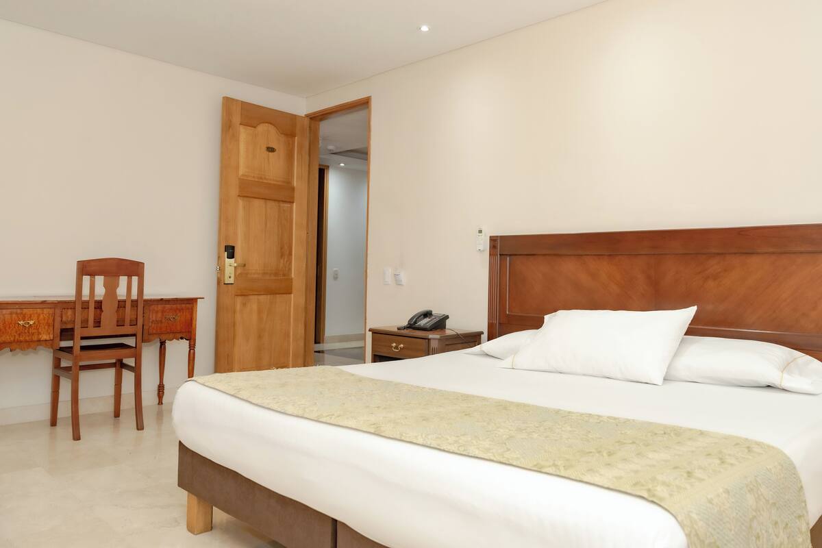 A hotel room with a wooden bed, a desk with a chair, and a bedside table with a phone.