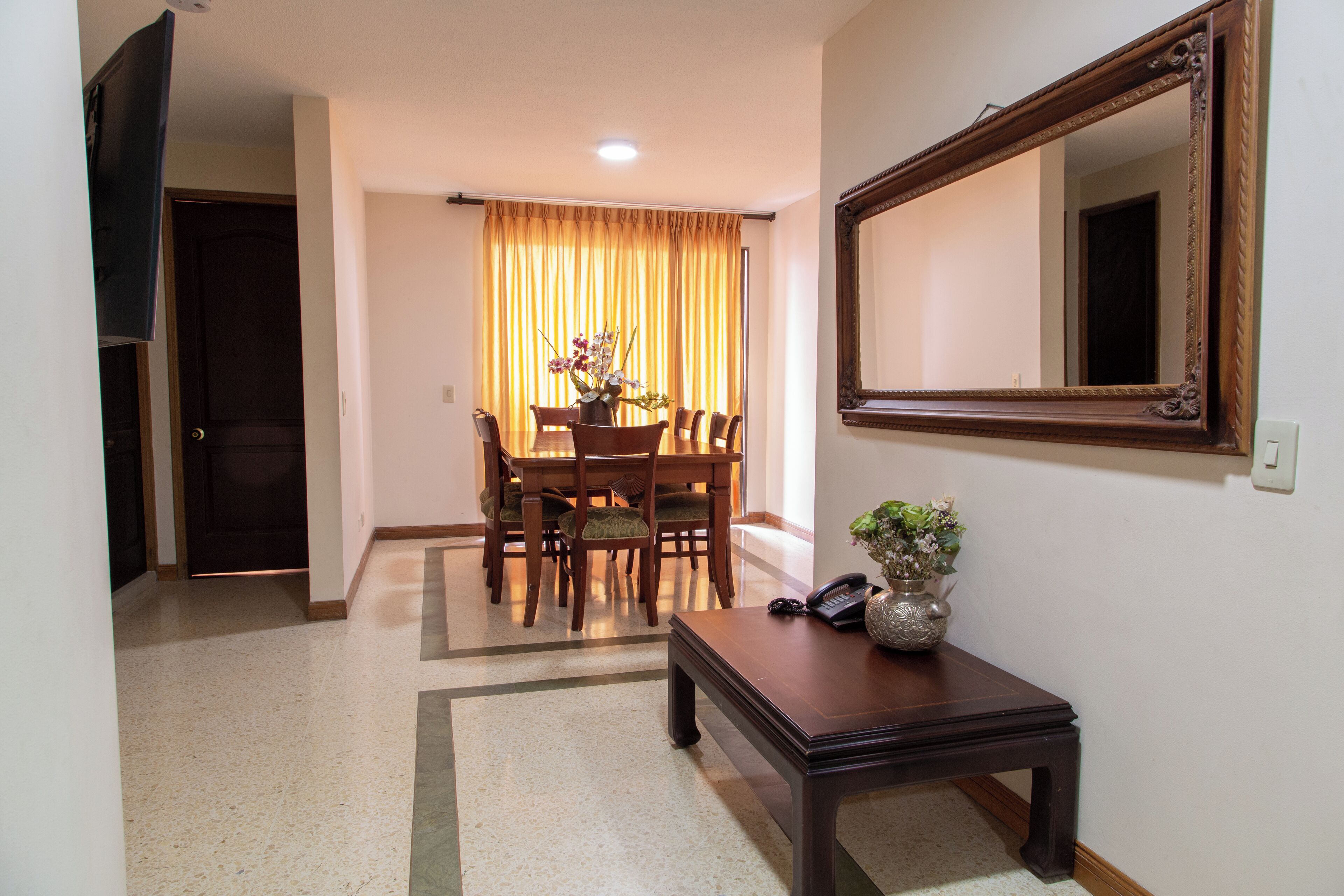 A dining area with a table and chairs, a vase with flowers, a telephone, and a mirror on the wall.