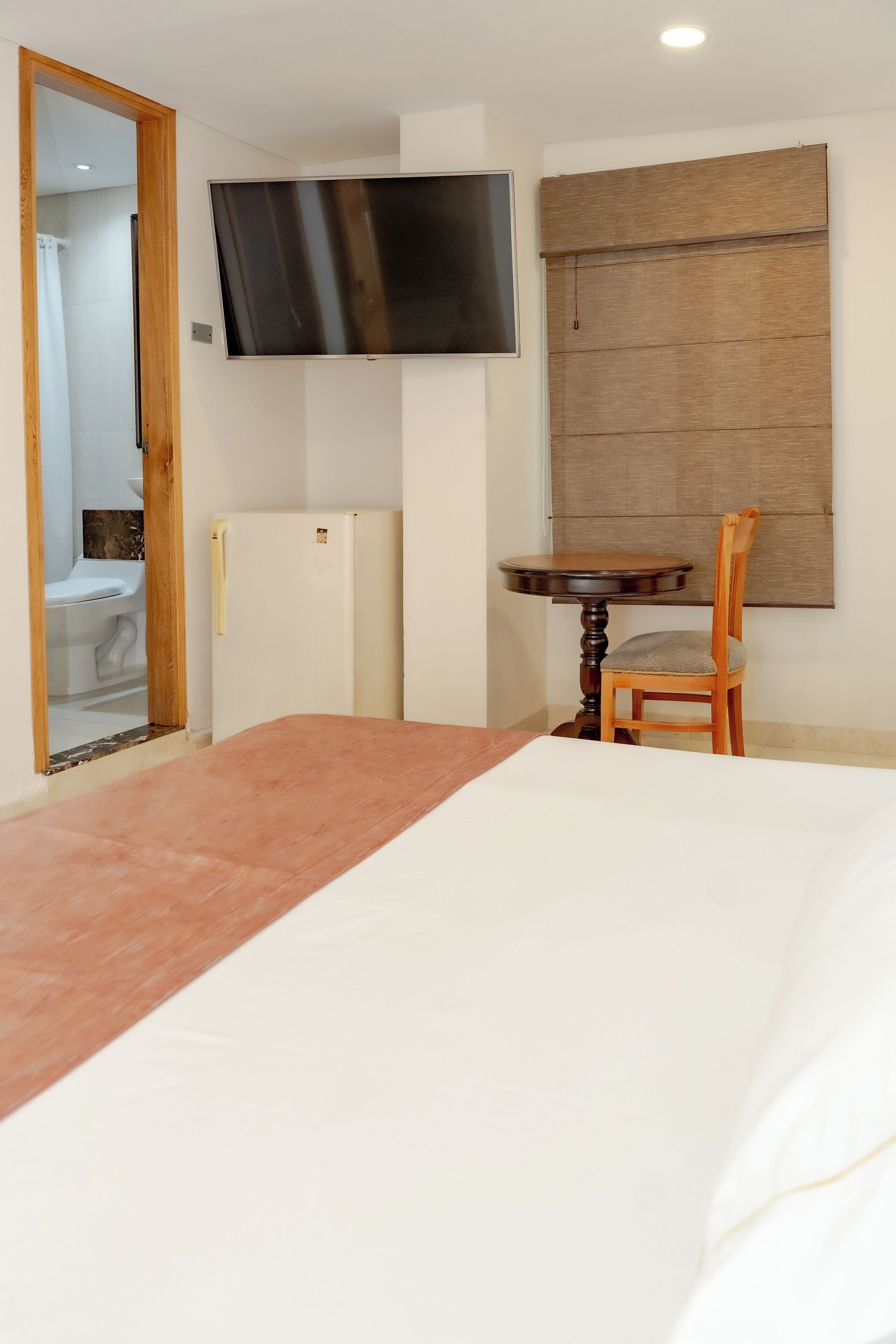 Een slaapkamer met een bed, een aan de muur bevestigde televisie, een klein tafeltje met een stoel en een koelkast.