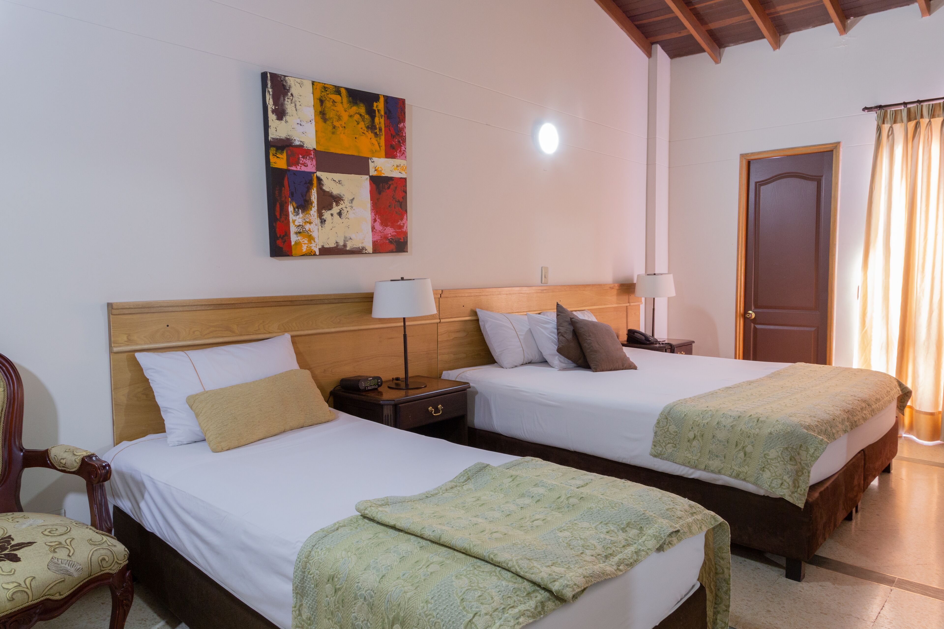 Een hotelkamer met twee bedden, een houten hoofdeinde, een nachtkastje met een lamp, een stoel en een kleurrijk abstract schilderij aan de muur.