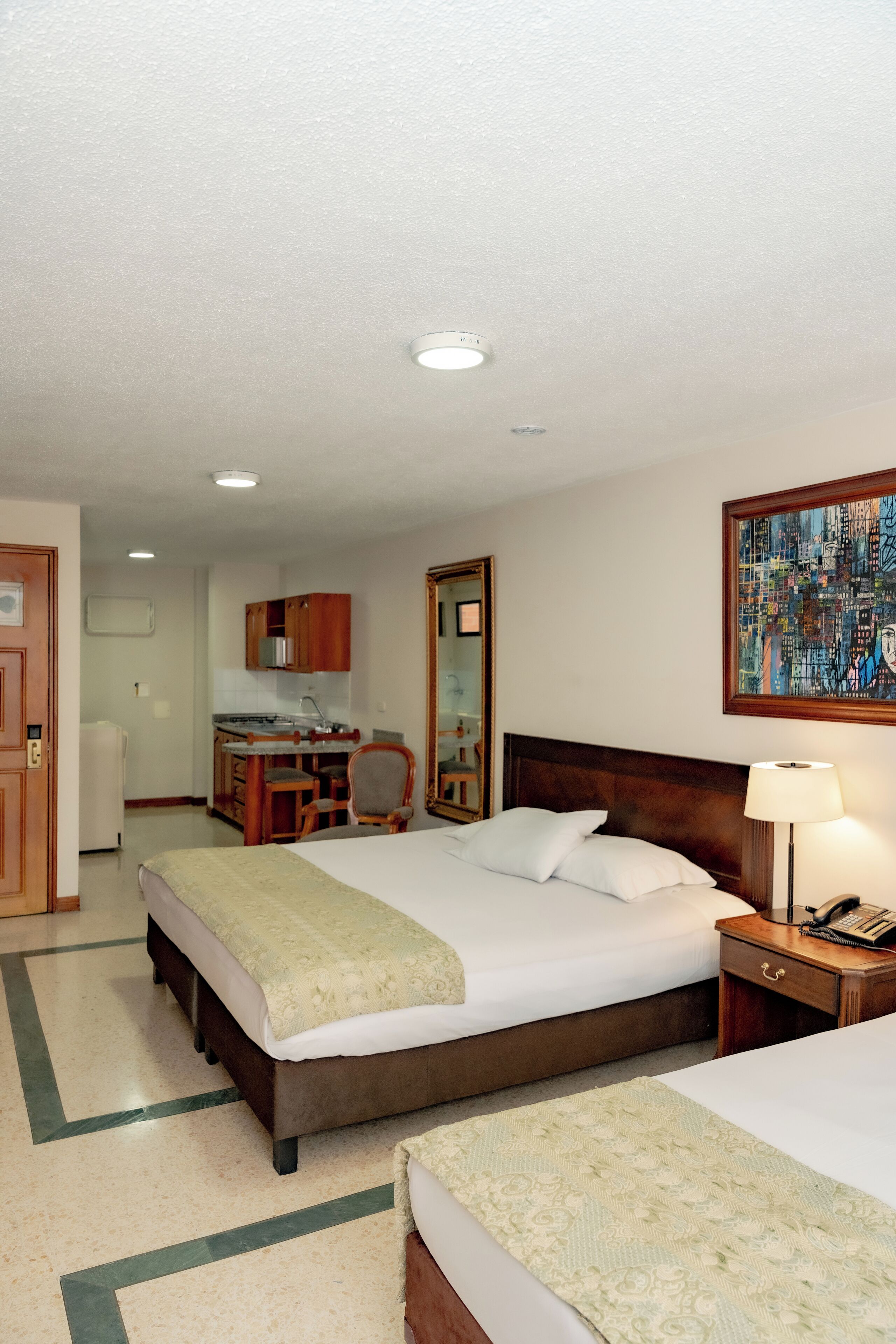 A hotel room with a large bed, a bedside table with a lamp, a desk with a chair, a mirror, and a painting on the wall.