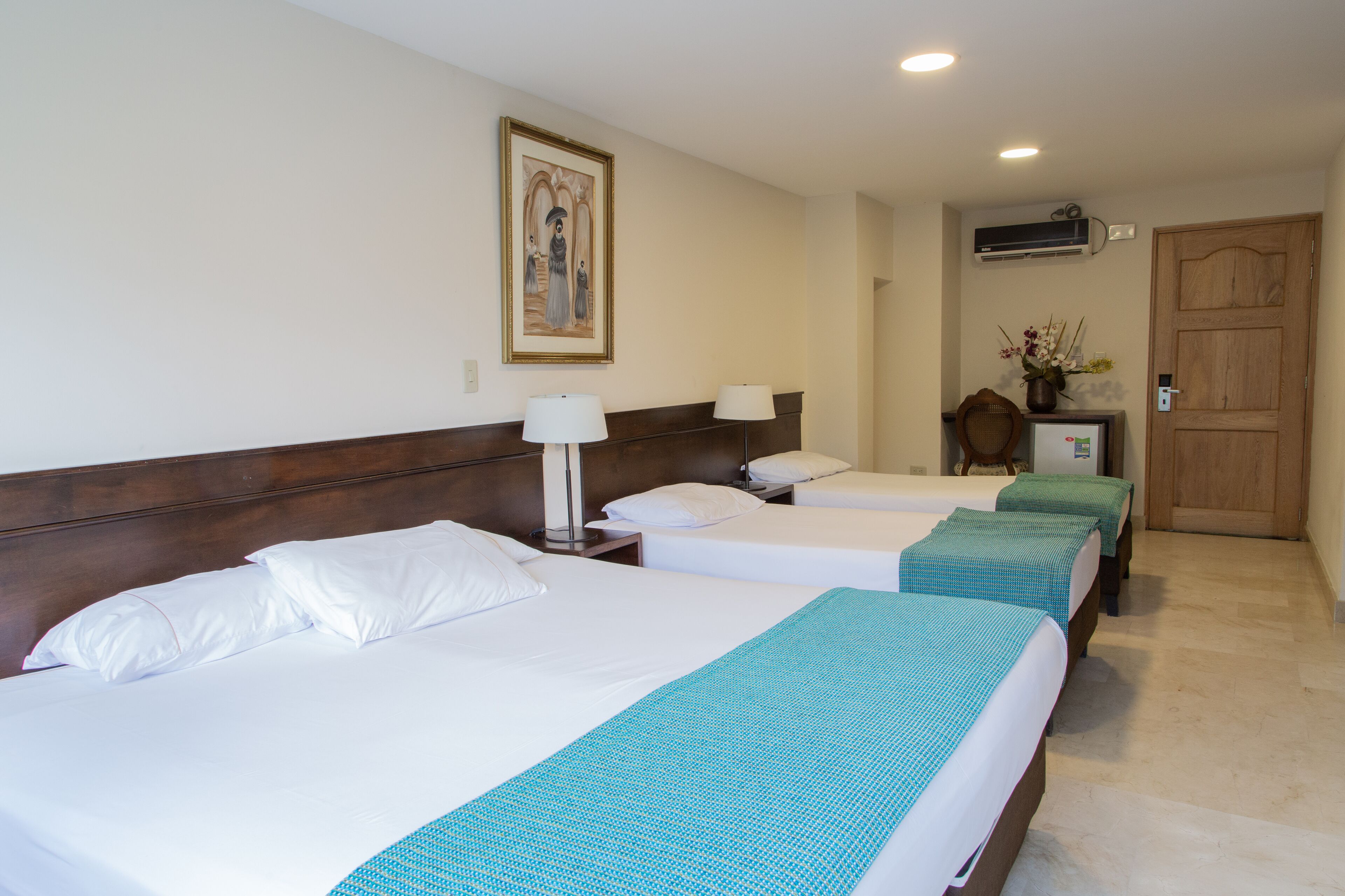 A hotel room with three beds, a wooden headboard, a painting on the wall, a wooden door, and an air conditioning unit.