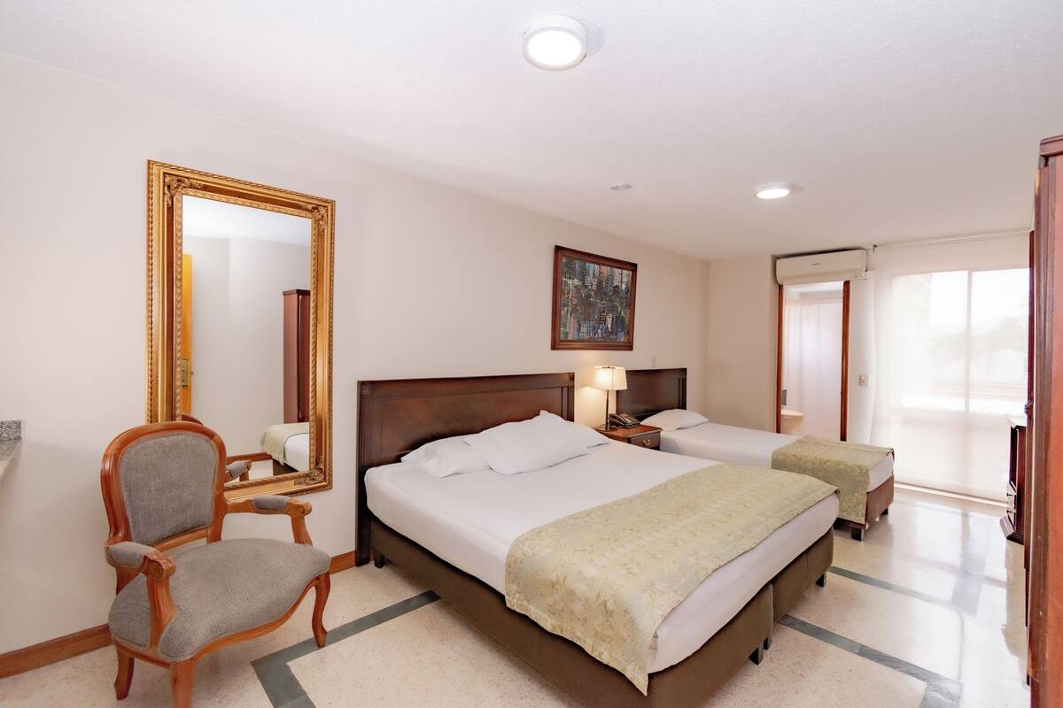A hotel room with two beds, a large mirror, a chair, a bedside table with a lamp, and a painting on the wall.