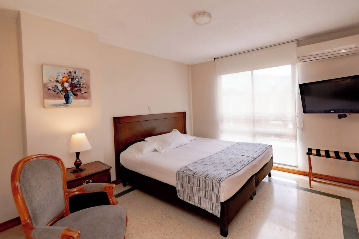 A hotel room with a bed, a chair, a painting, a TV, and a window.