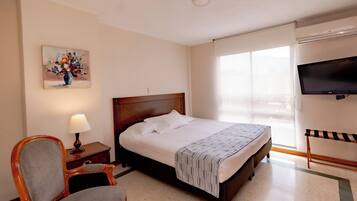 A hotel room with a bed, a chair, a painting, a TV, and a window.