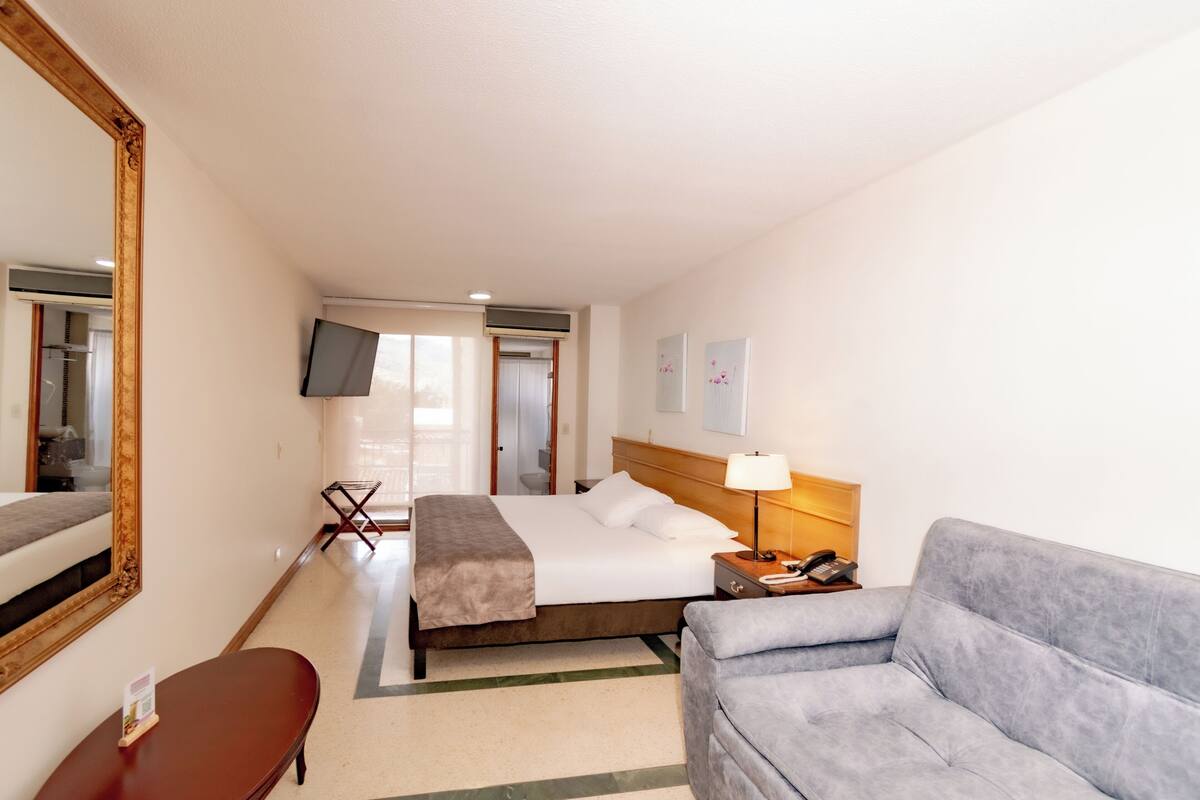 A hotel room with a bed, a sofa, a desk, a mirror, a TV, and a window.