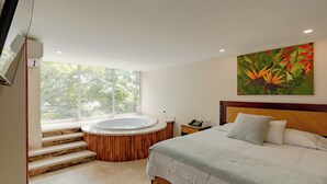 Un dormitorio moderno con una cama grande, cabecera de madera, un cuadro de flores tropicales, una bañera integrada y una ventana con vistas a árboles.