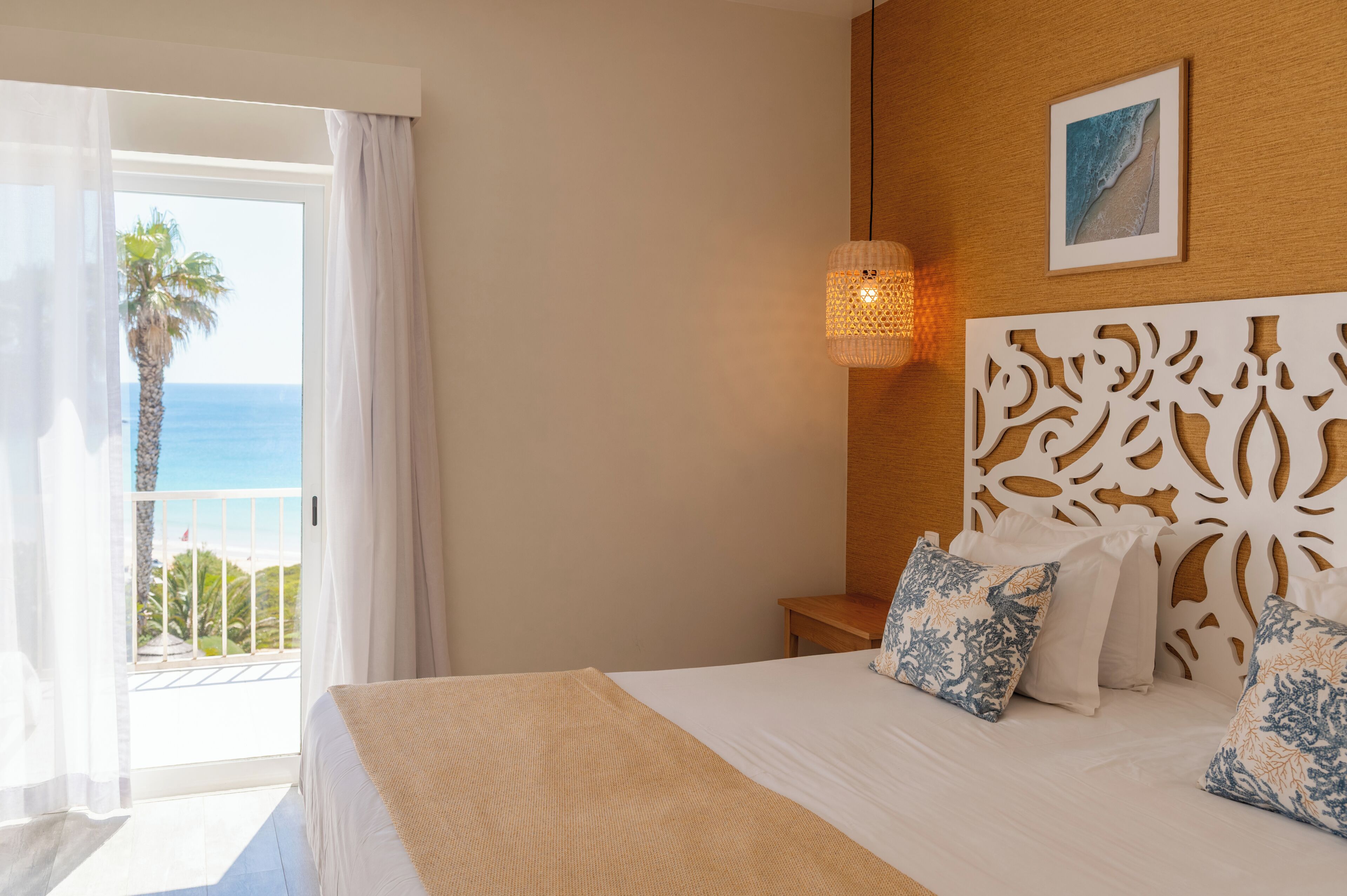 A hotel room with a bed, a nightstand, a decorative headboard, a wall-mounted picture, and a view of the ocean through a balcony.