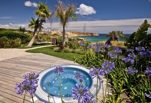 Una terraza de madera con un jacuzzi rodeado de flores coloridas, con vistas a un paisaje costero con palmeras y cielo despejado.