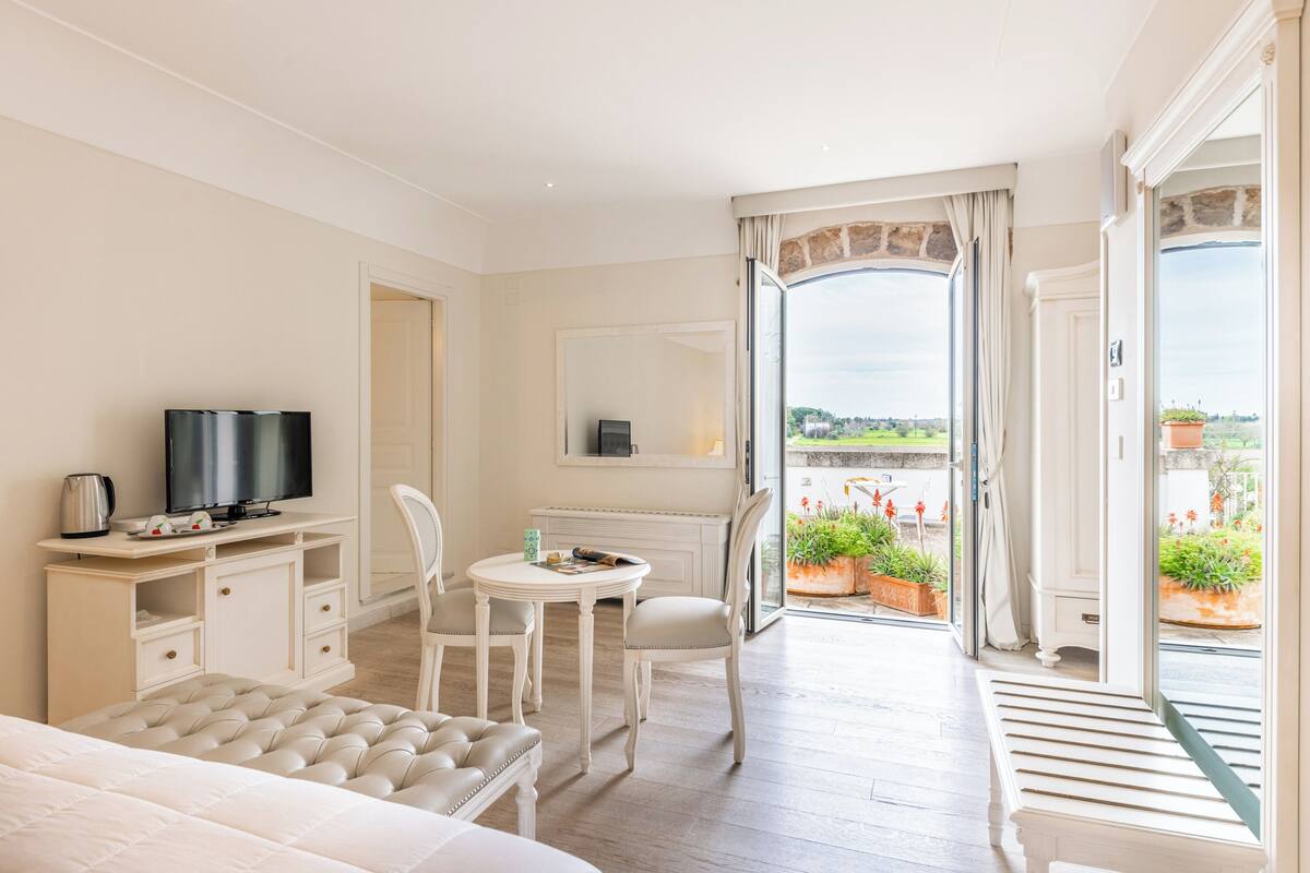 A hotel room with a bed, a TV, a desk with a computer, a chair, a small table, and a balcony with plants.