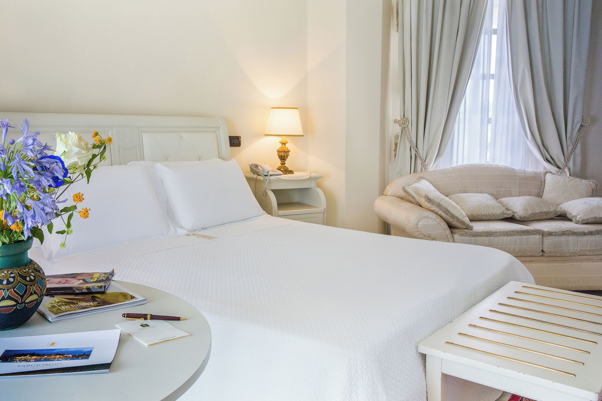 Een hotelkamer met een bed, nachtkastje, lamp, stoel, en een klein tafeltje met een bloemenvaas en enkele voorwerpen.