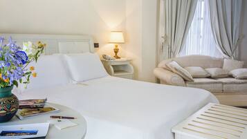 Een hotelkamer met een bed, nachtkastje, lamp, stoel, en een klein tafeltje met een bloemenvaas en enkele voorwerpen.
