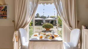 A dining table with breakfast items, two white chairs, and a view of a garden and gazebo through a window with curtains. - Sangiorgio Resort & Spa (Cutrofiano)