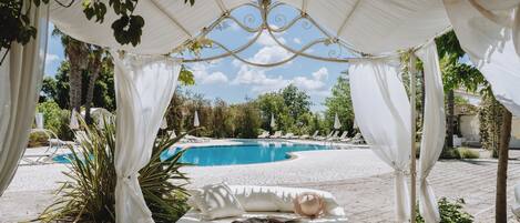 Una cama con dosel, una piscina y sillones de descanso al fondo.