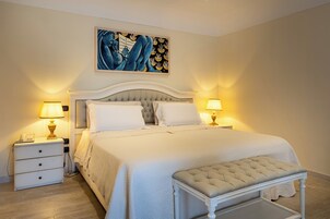 Un dormitorio con cama, lámparas de noche, mesitas de noche, un banco y un cuadro en la pared.