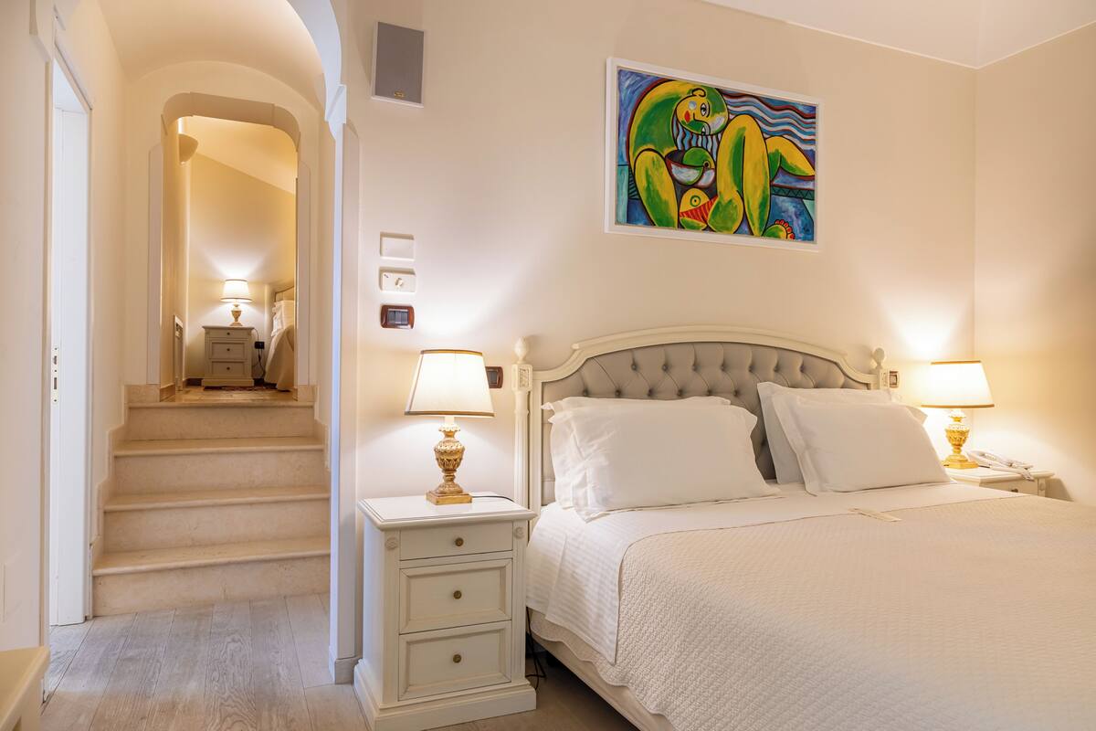 A bedroom with a bed, nightstands, a lamp, a painting, and a staircase leading to another room.