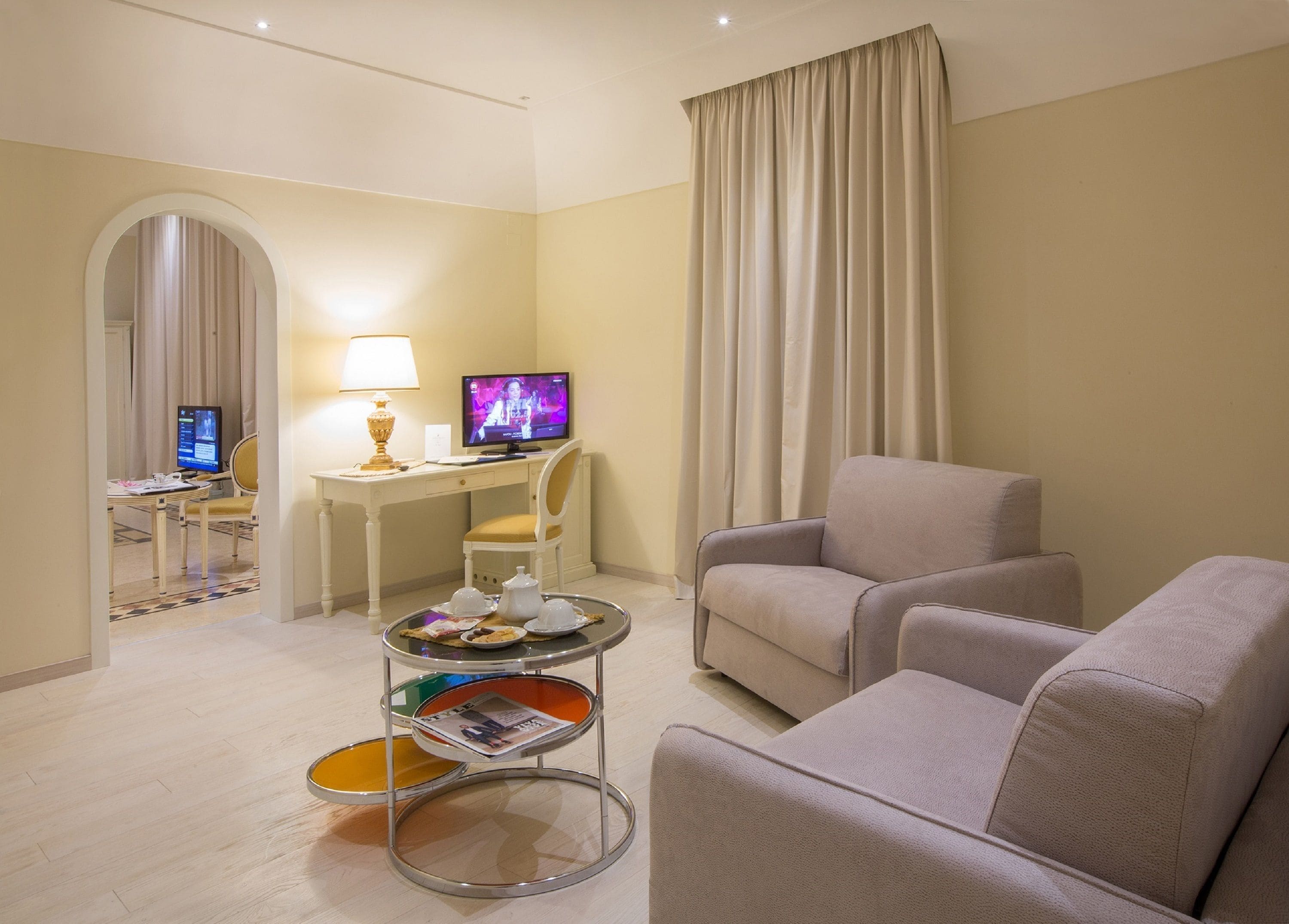 A hotel room with a seating area, a desk with a computer, a television, and a small table with magazines.