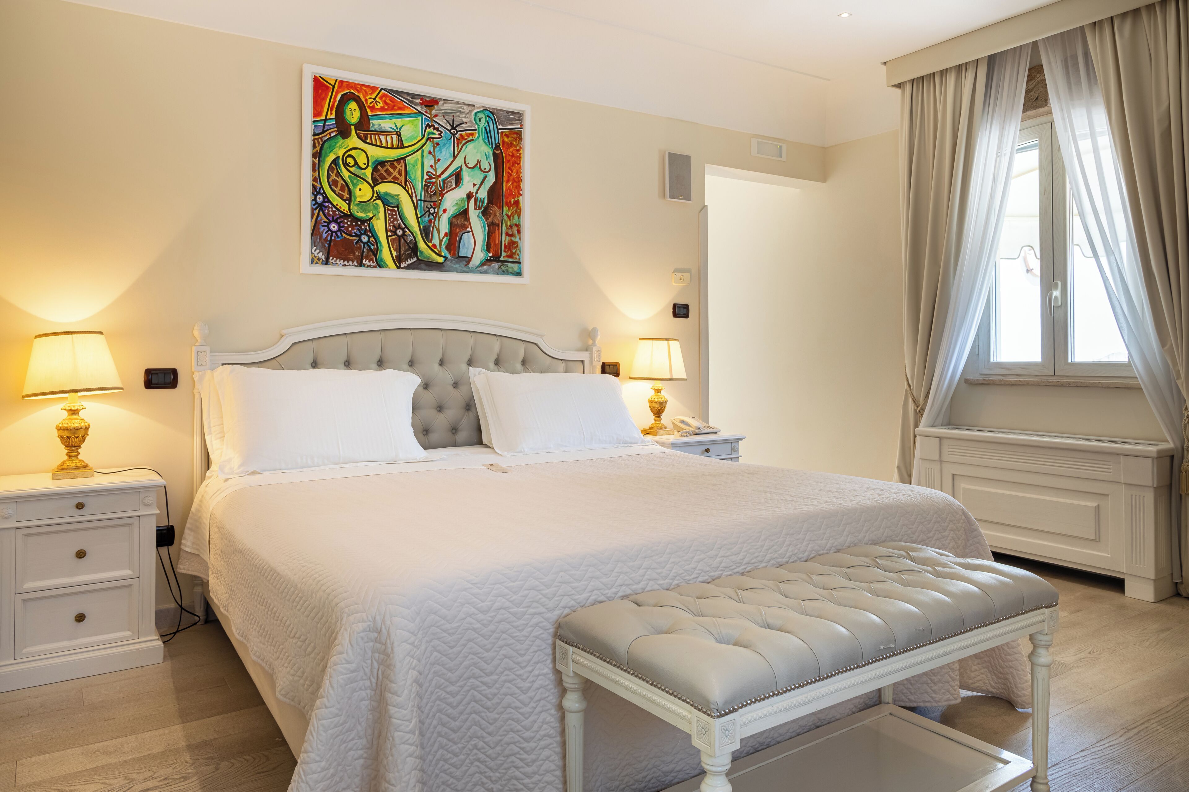 Un dormitorio con cama, mesitas de noche, un banco, un cuadro y una ventana con cortinas.