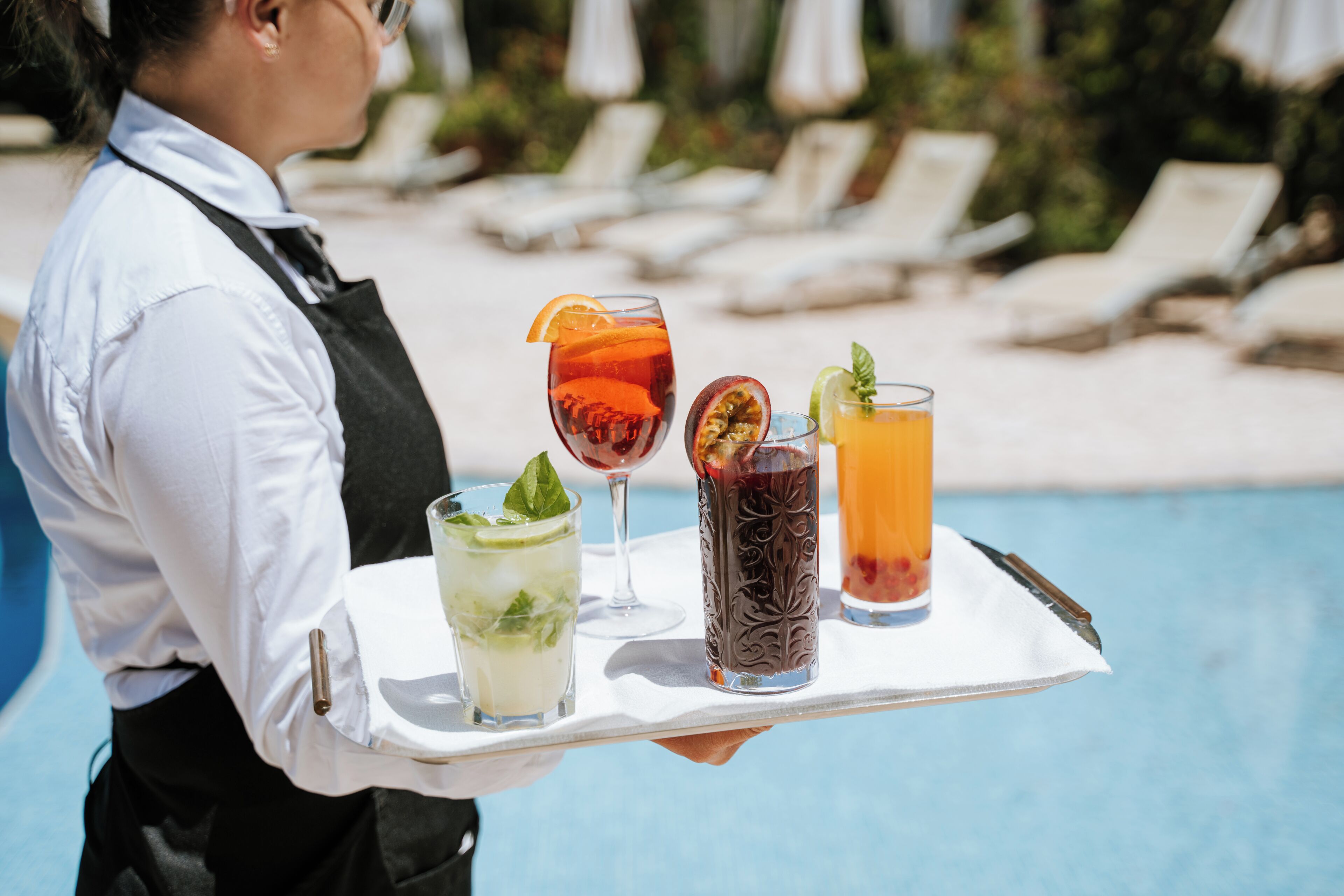 A person holding a tray with various drinks, a pool, and lounge chairs in the background.