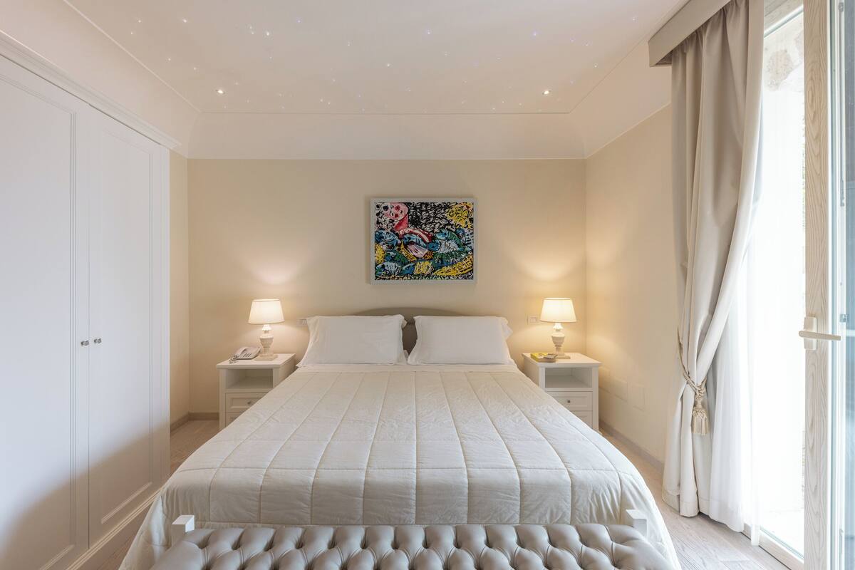 A bedroom with a bed, two bedside tables with lamps, a wall-mounted artwork, and a large window with curtains.