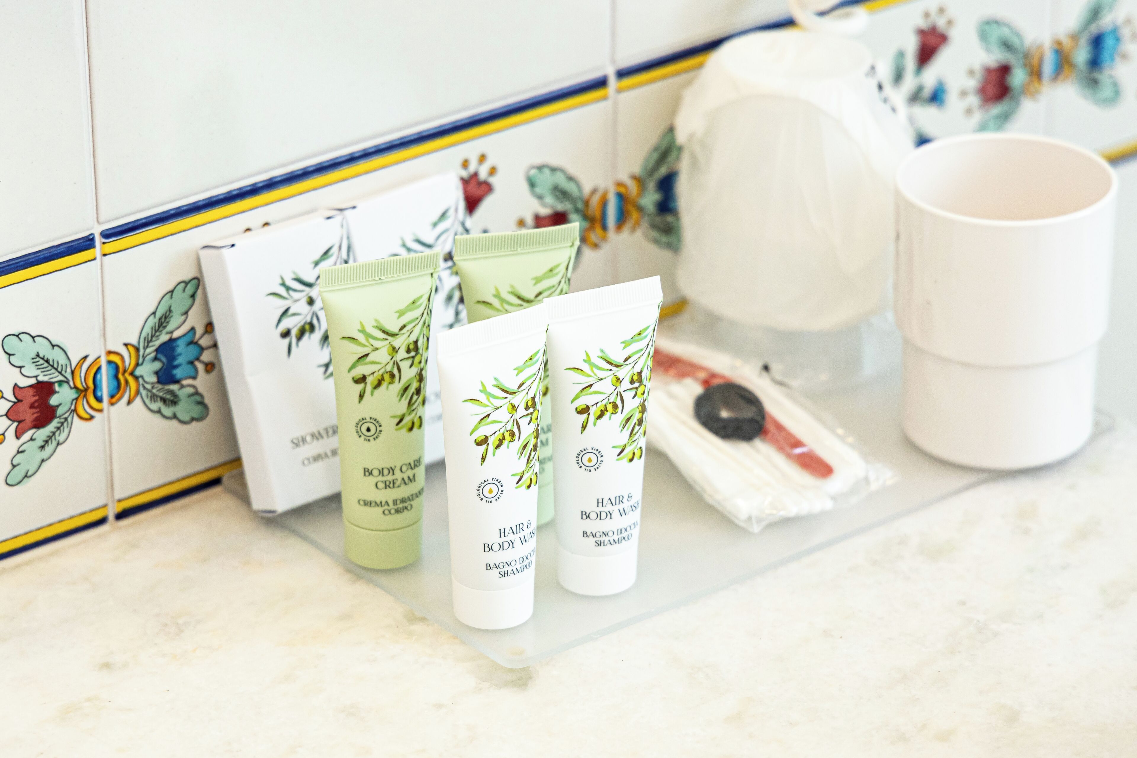 A bathroom with a tiled backsplash, a white sink, and toiletries including a shampoo bottle, a body cream tube, and a cup.