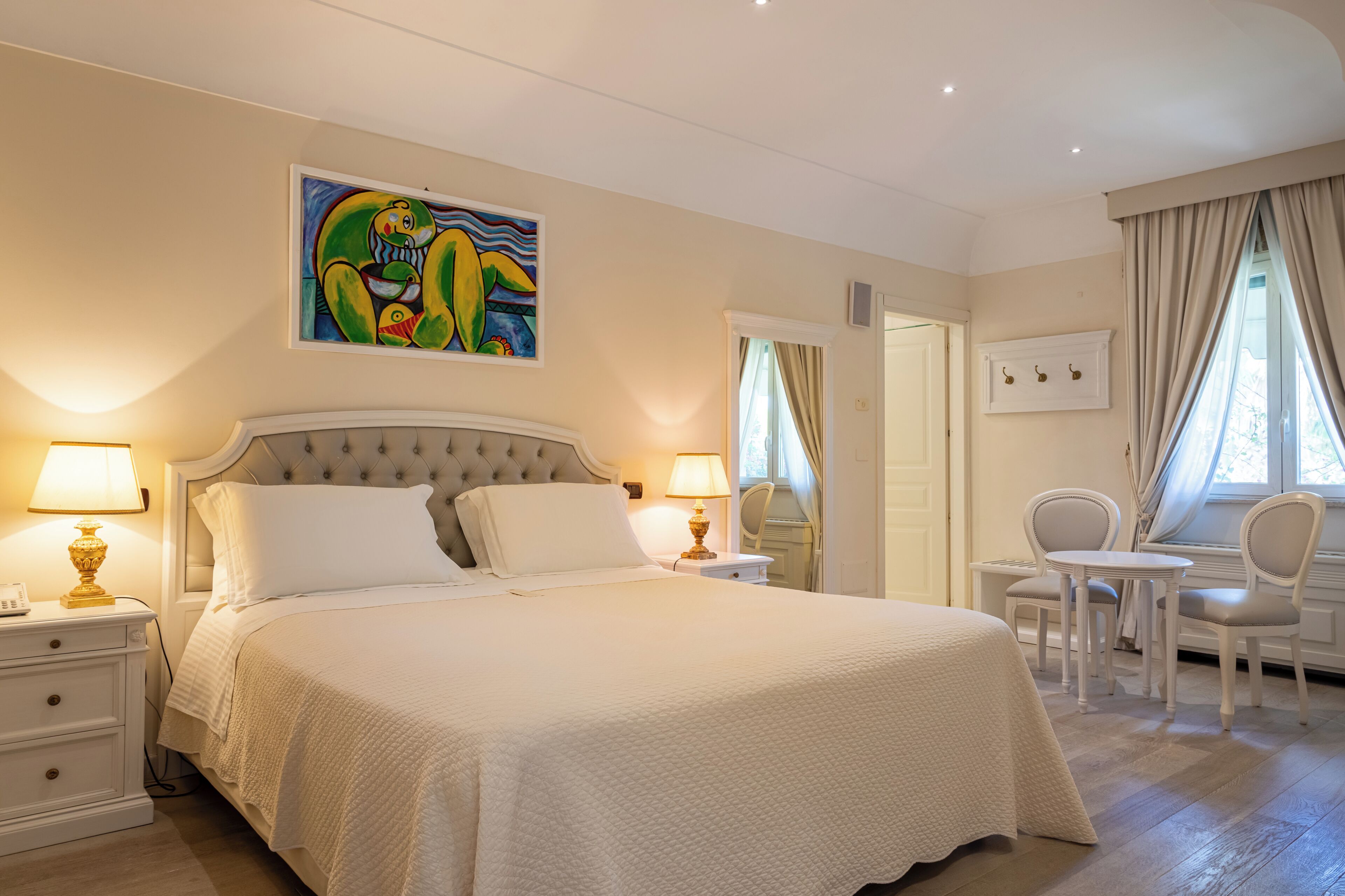 A bedroom with a large bed, bedside lamps, a painting, a desk with chairs, and a door leading to another room.