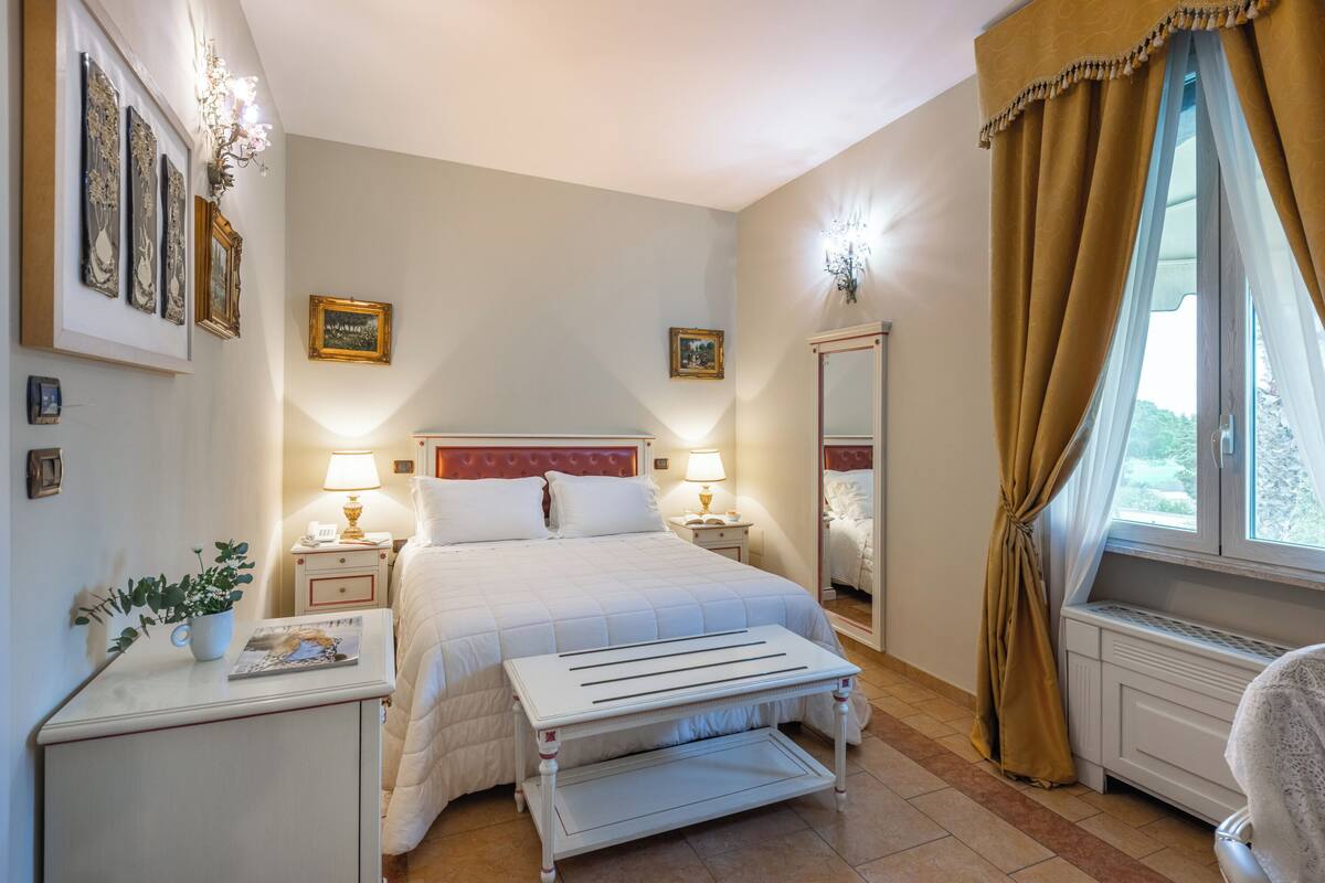 A neatly arranged bedroom with a bed, bedside tables, a nightstand, a mirror, and a window with curtains.