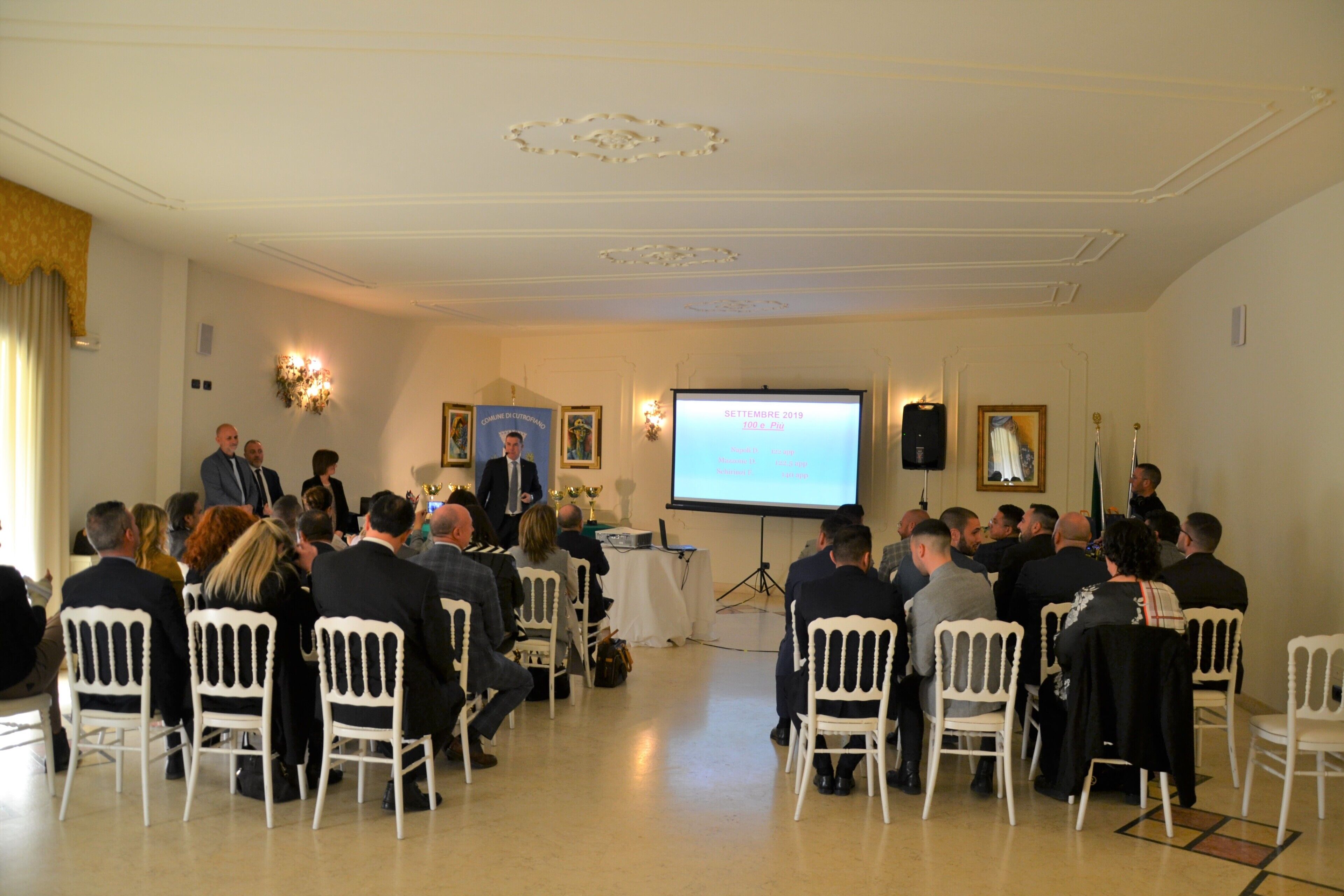 A formal meeting room with white chairs arranged in rows, a podium, and a presentation screen.