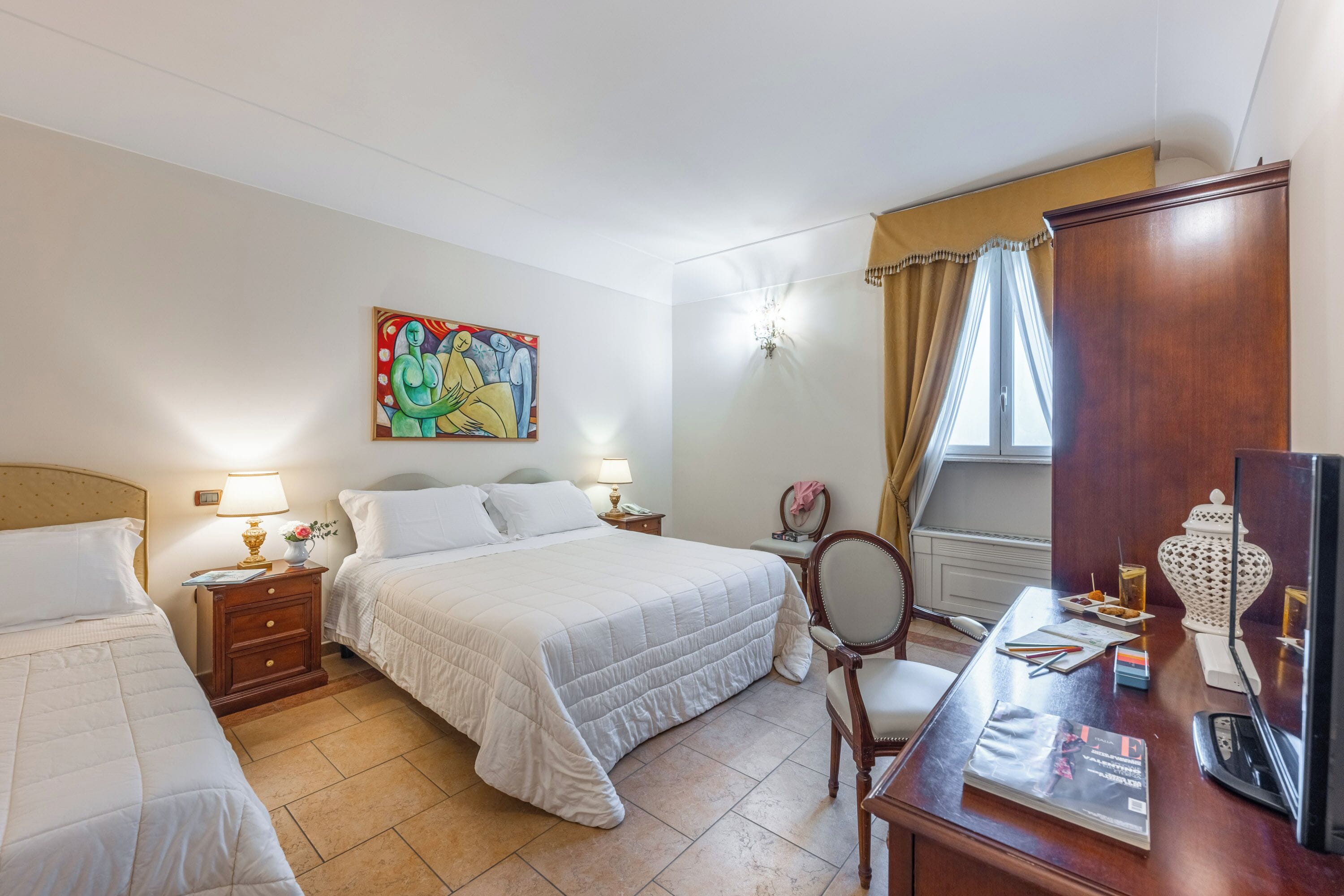 A hotel room with two beds, a desk with a chair, a TV, a lamp, a painting, and a window with curtains.