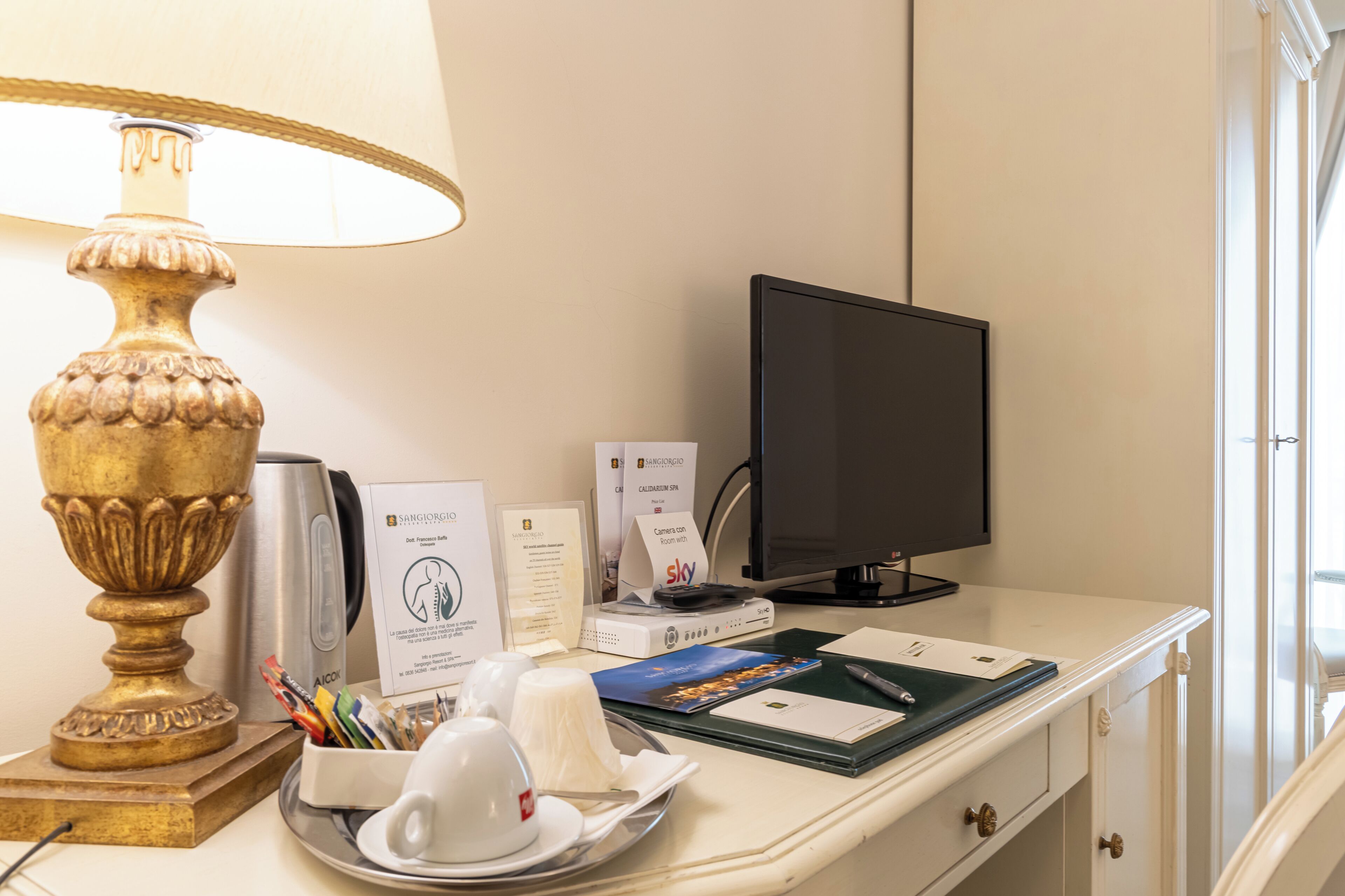 A hotel room desk with a lamp, a computer monitor, a coffee cup, a plate with a napkin, and some documents.
