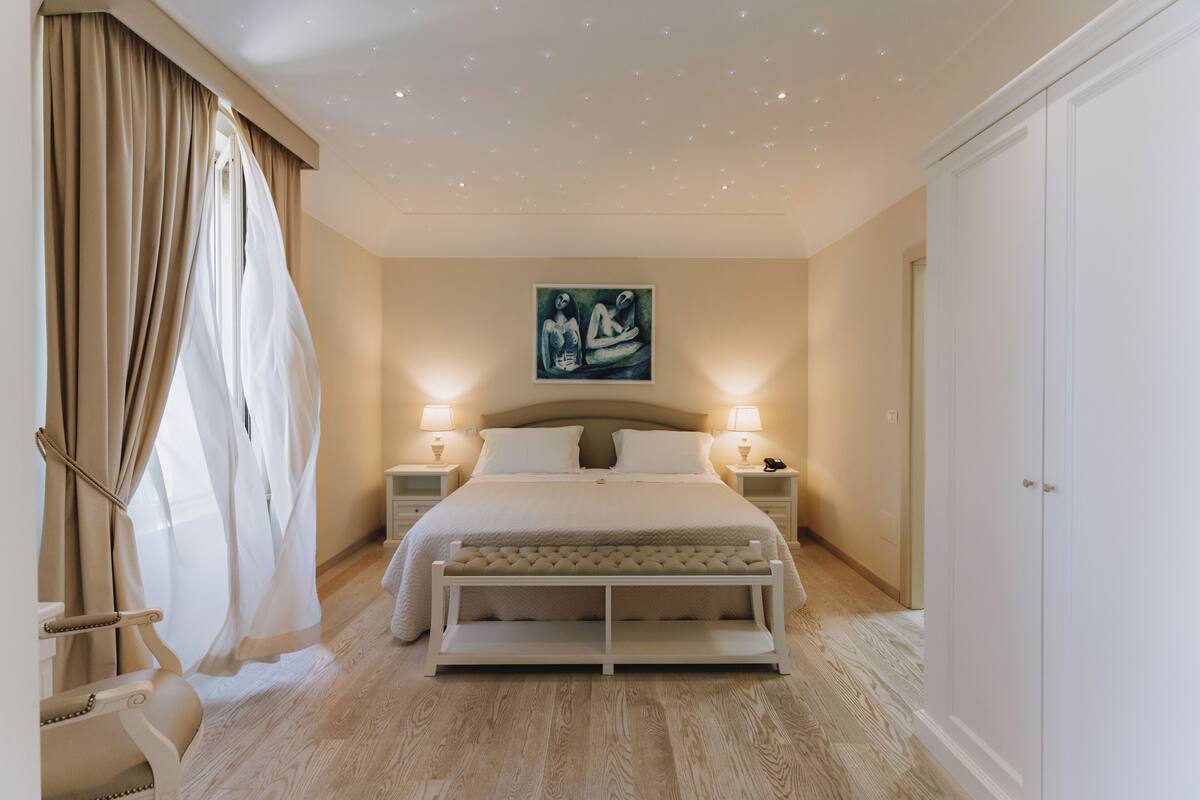 A bedroom with a bed, bedside tables, a bench, a painting, and a door.