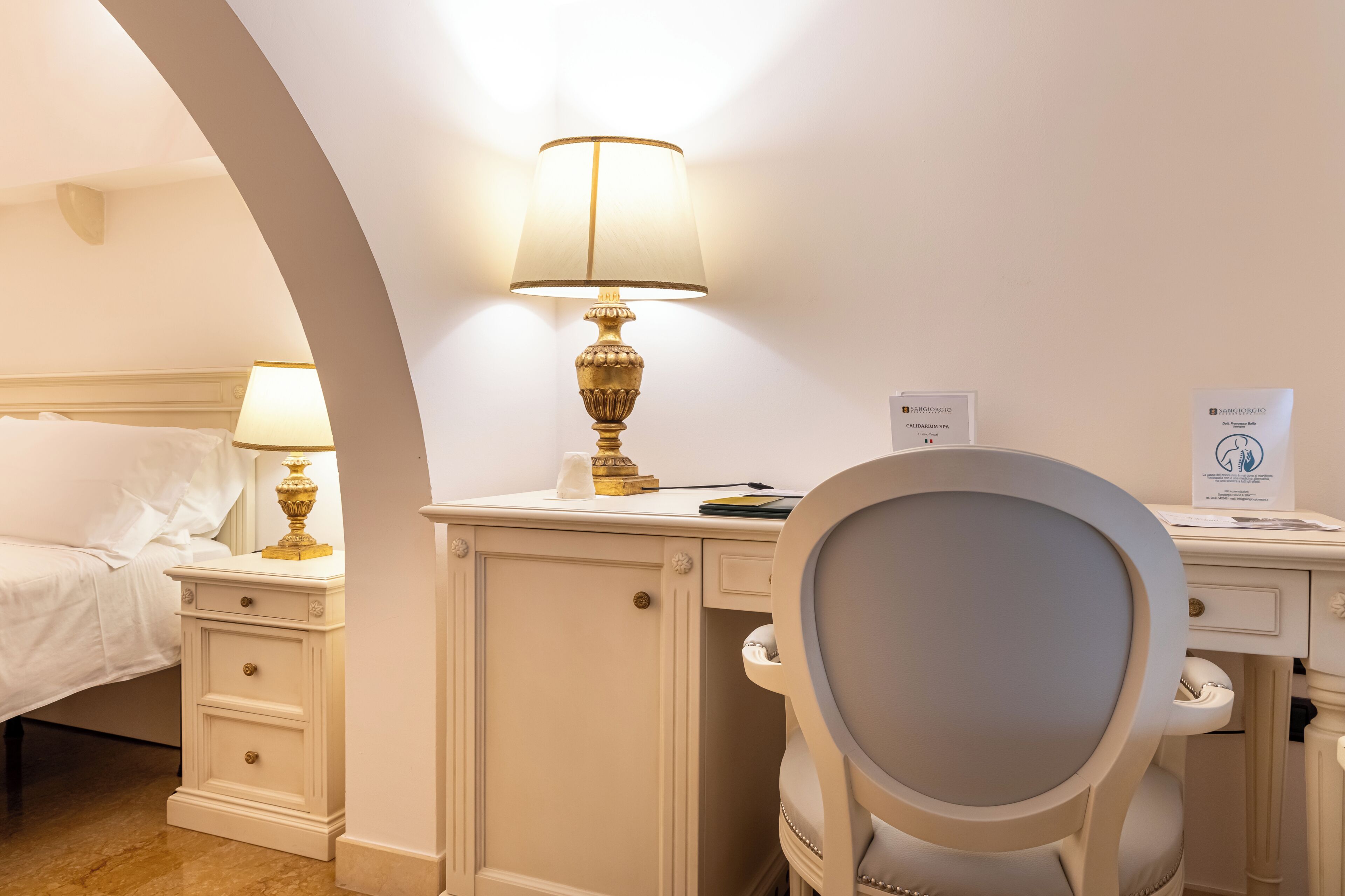 A hotel room with a bed, a desk, a chair, a lamp, and a sign indicating a spa service.