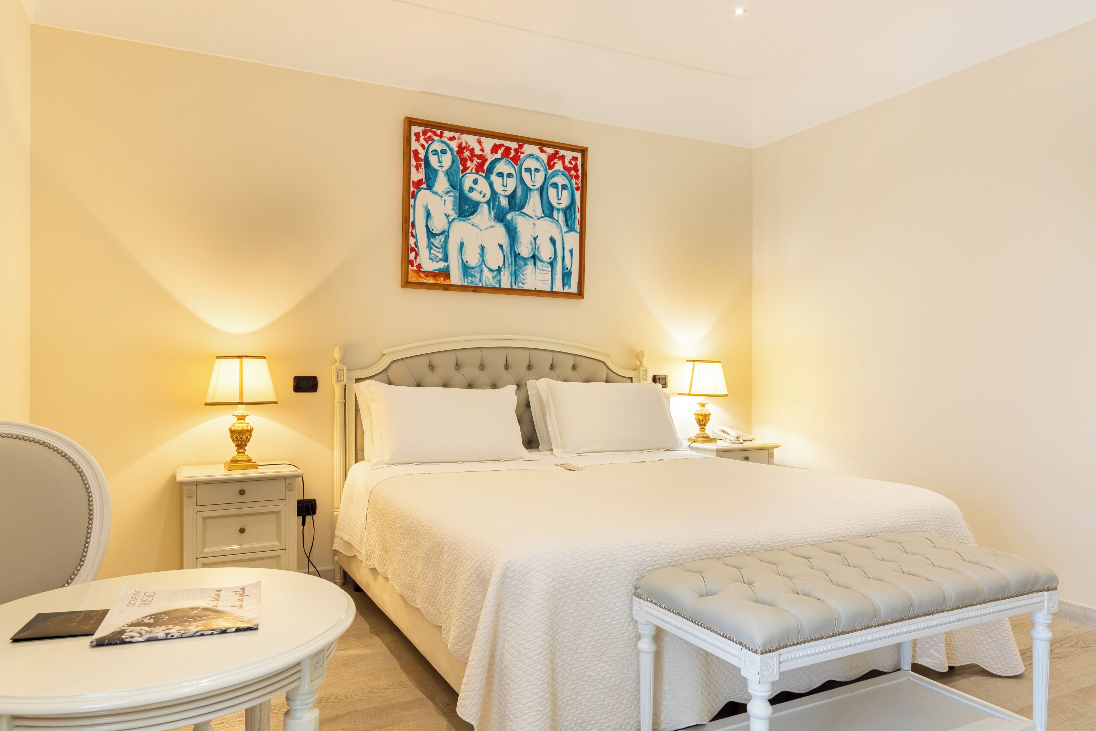 A bedroom with a bed, bedside lamps, a chair, a table, and a painting on the wall.