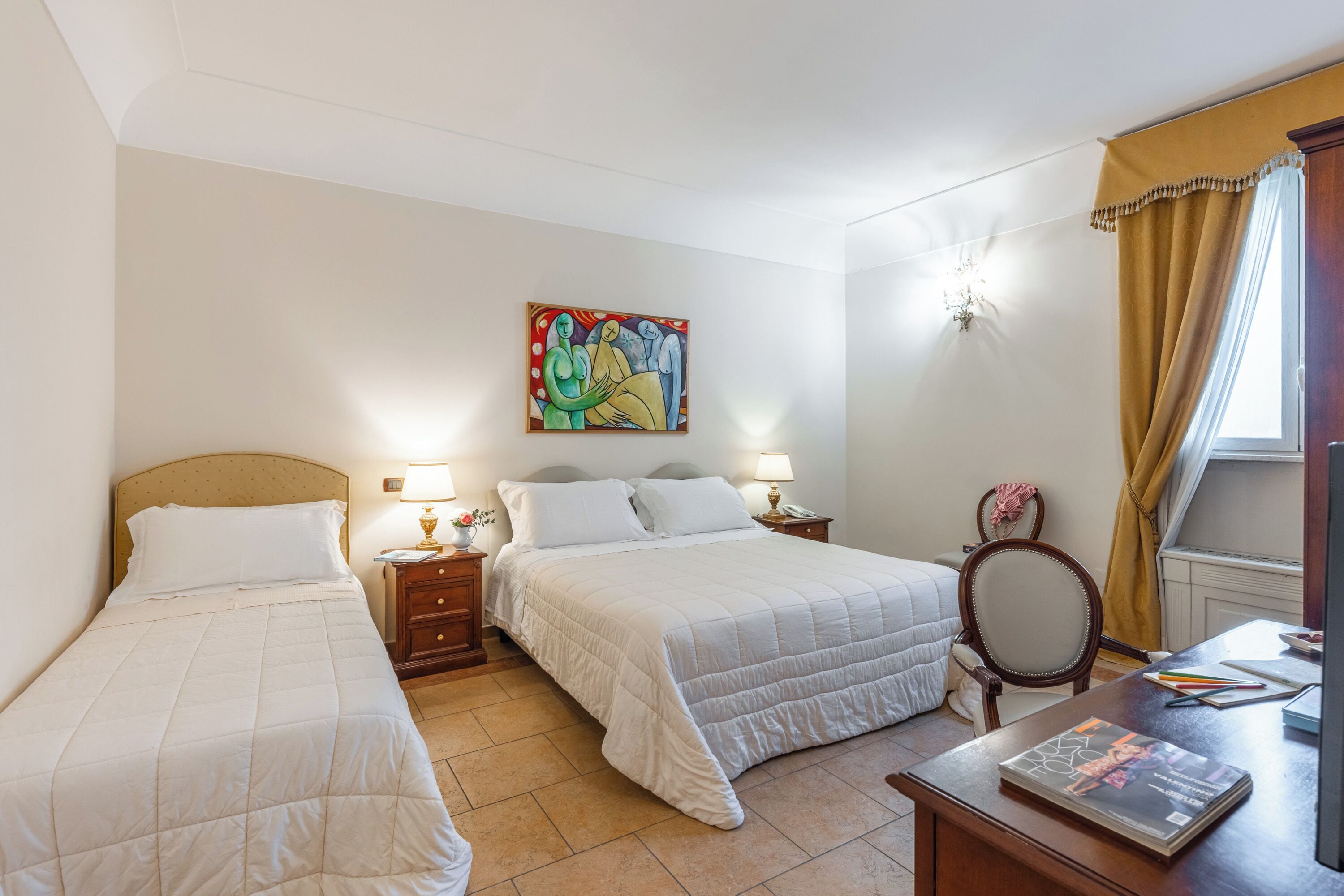 A hotel room with two beds, a painting on the wall, a desk with a magazine, and a window with curtains.