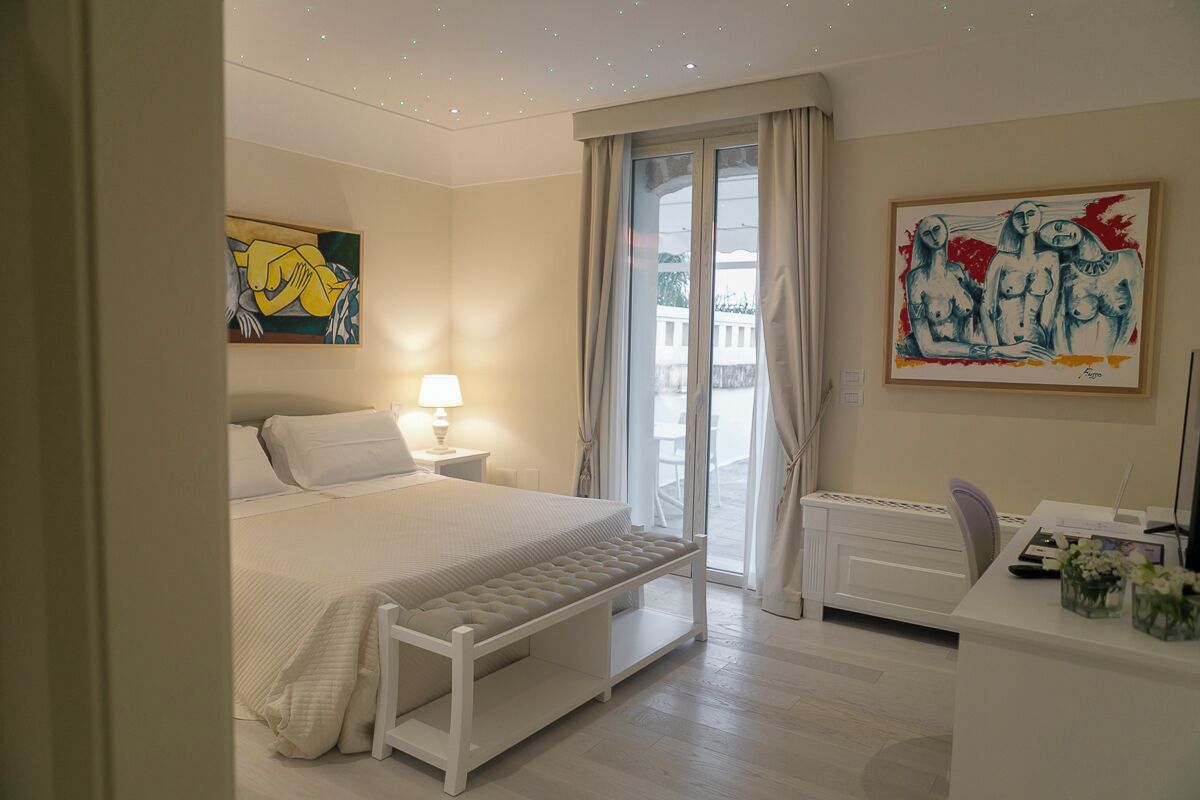 A hotel room with a bed, bedside table, lamp, desk, chair, and artwork on the walls.