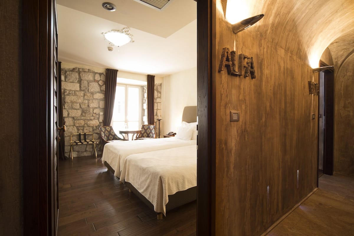 Une chambre d’hôtel avec un mur en pierre, un lit recouvert d’une courtepointe blanche, une porte en bois avec une plaque nominative et une petite table avec des chaises.