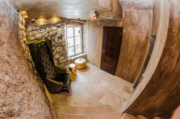 Una habitación con paredes de piedra, una puerta de madera, una ventana, un sillón verde y una mesa pequeña con un cuenco.