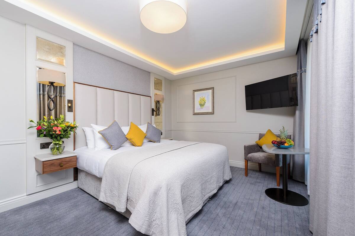 A modern hotel room with a large bed, bedside table, and a small round table with fruit.