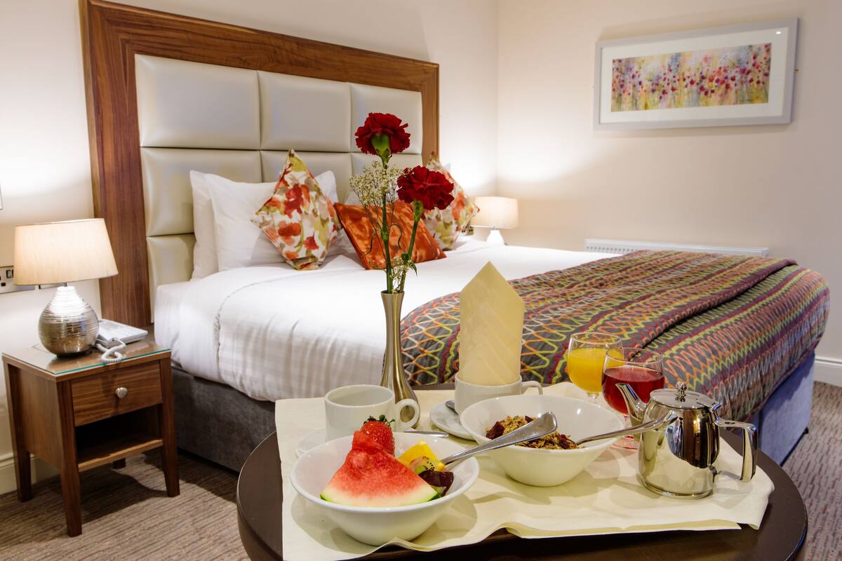A neatly made bed with a patterned bedspread, a bedside table with a lamp, and a tray with breakfast items including fruit, cereal, and a teapot.